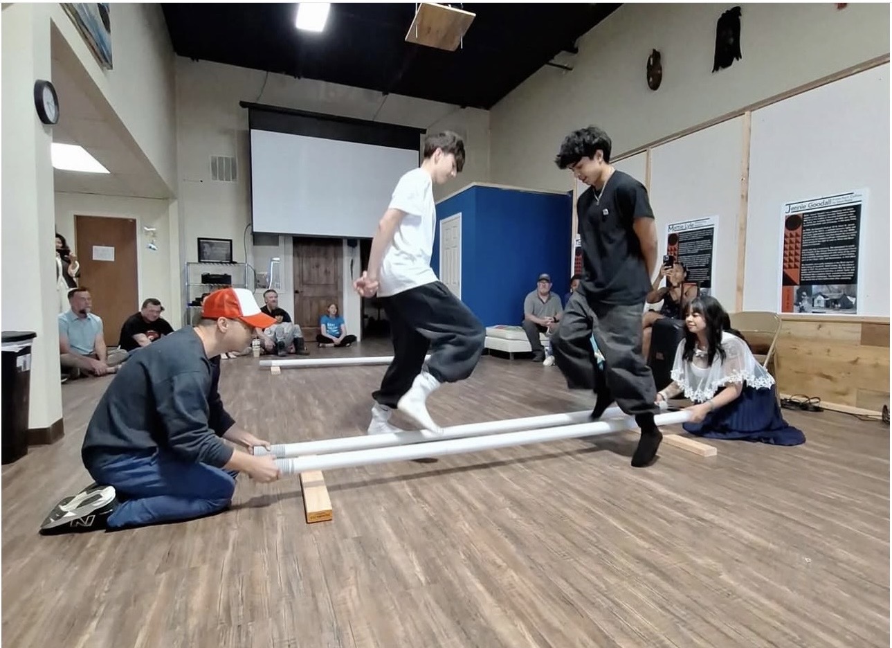 Group of people in a room with wooden floor, some sitting, others standing, two people balancing on a pipe, others assisting.