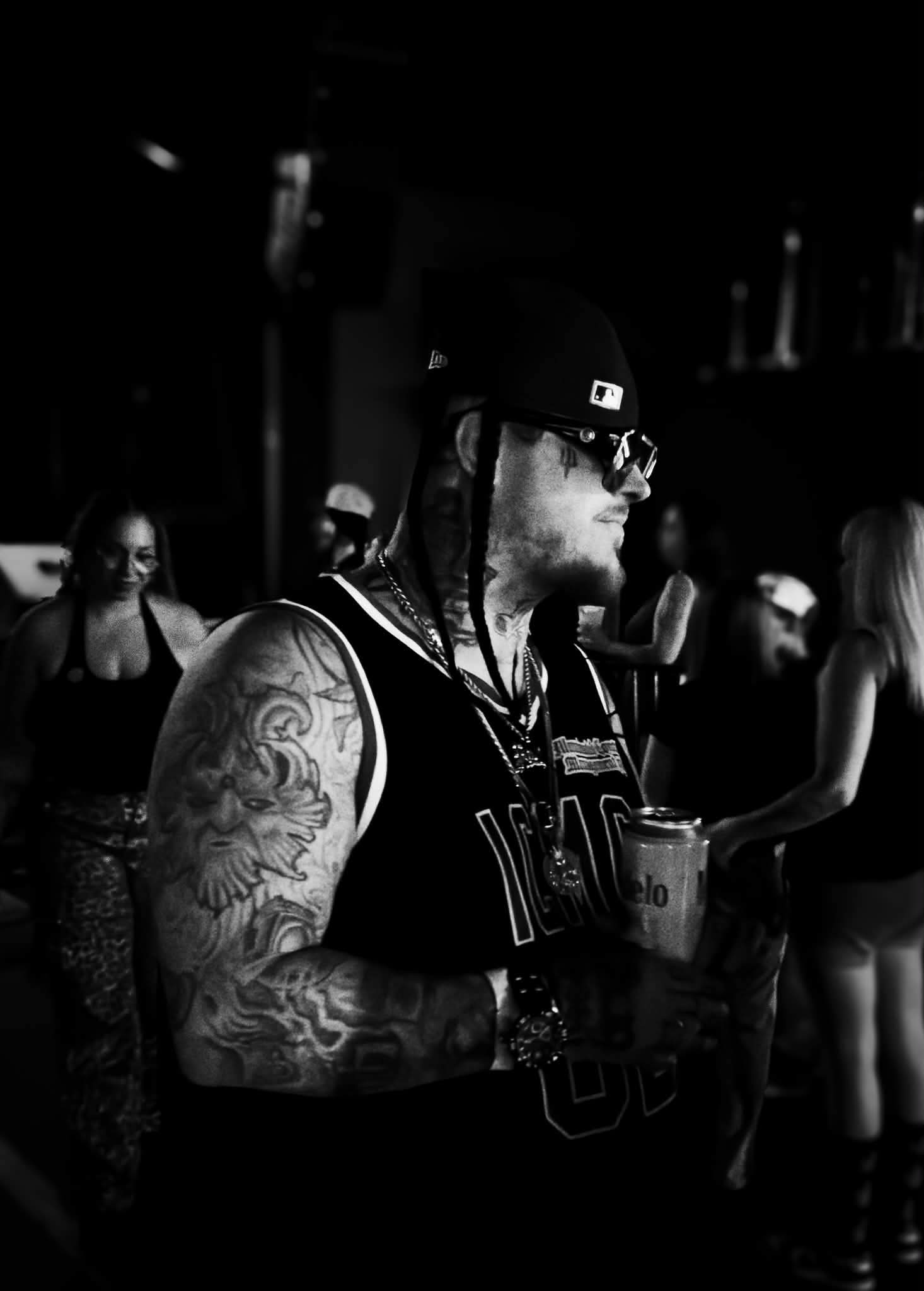 Man with tattoos, sunglasses, and a cap holding a drink in a crowded indoor setting.
