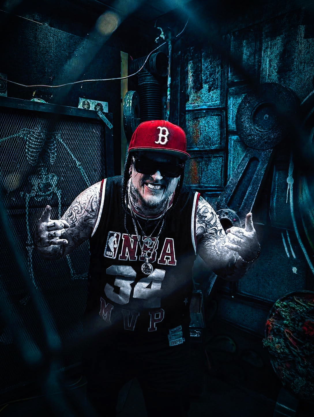 Person wearing a red cap, sunglasses, and a sleeveless shirt, smiling and giving two middle fingers, in a dark room with equipment.