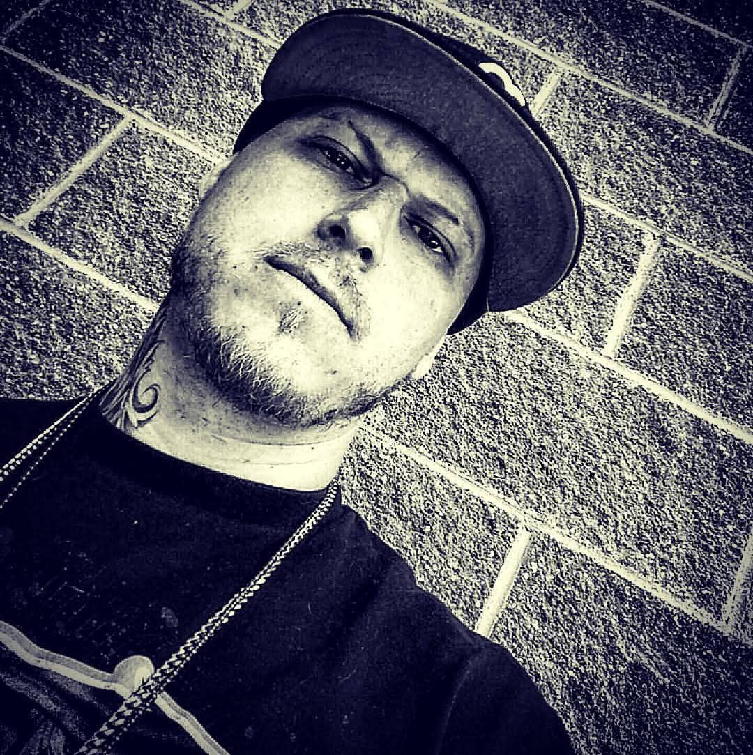 Man wearing a cap and chain, leaning against a brick wall, looking at the camera, black and white photo.