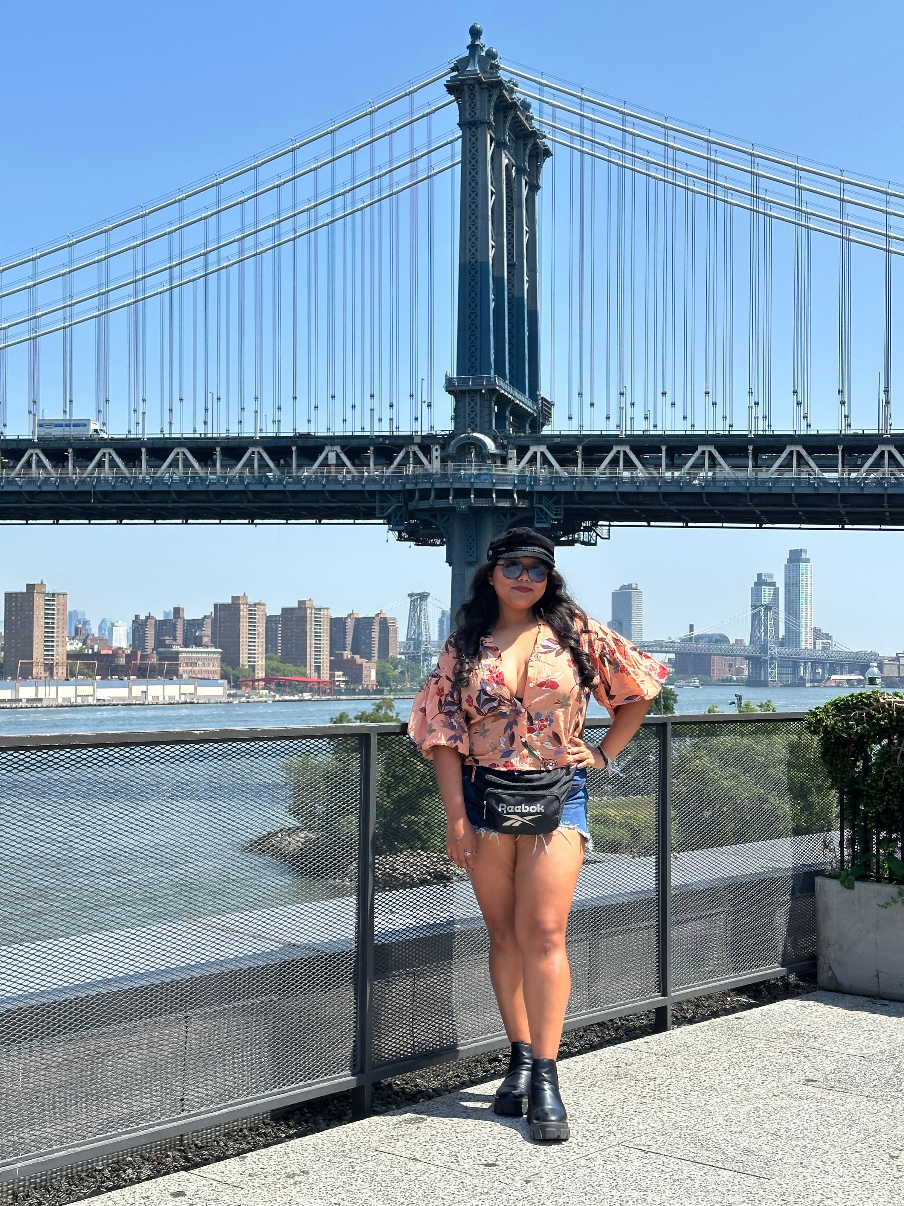 Woman standing on a bridge with a city skyline and river in the background, holding a phone, wearing a floral top and shorts.
