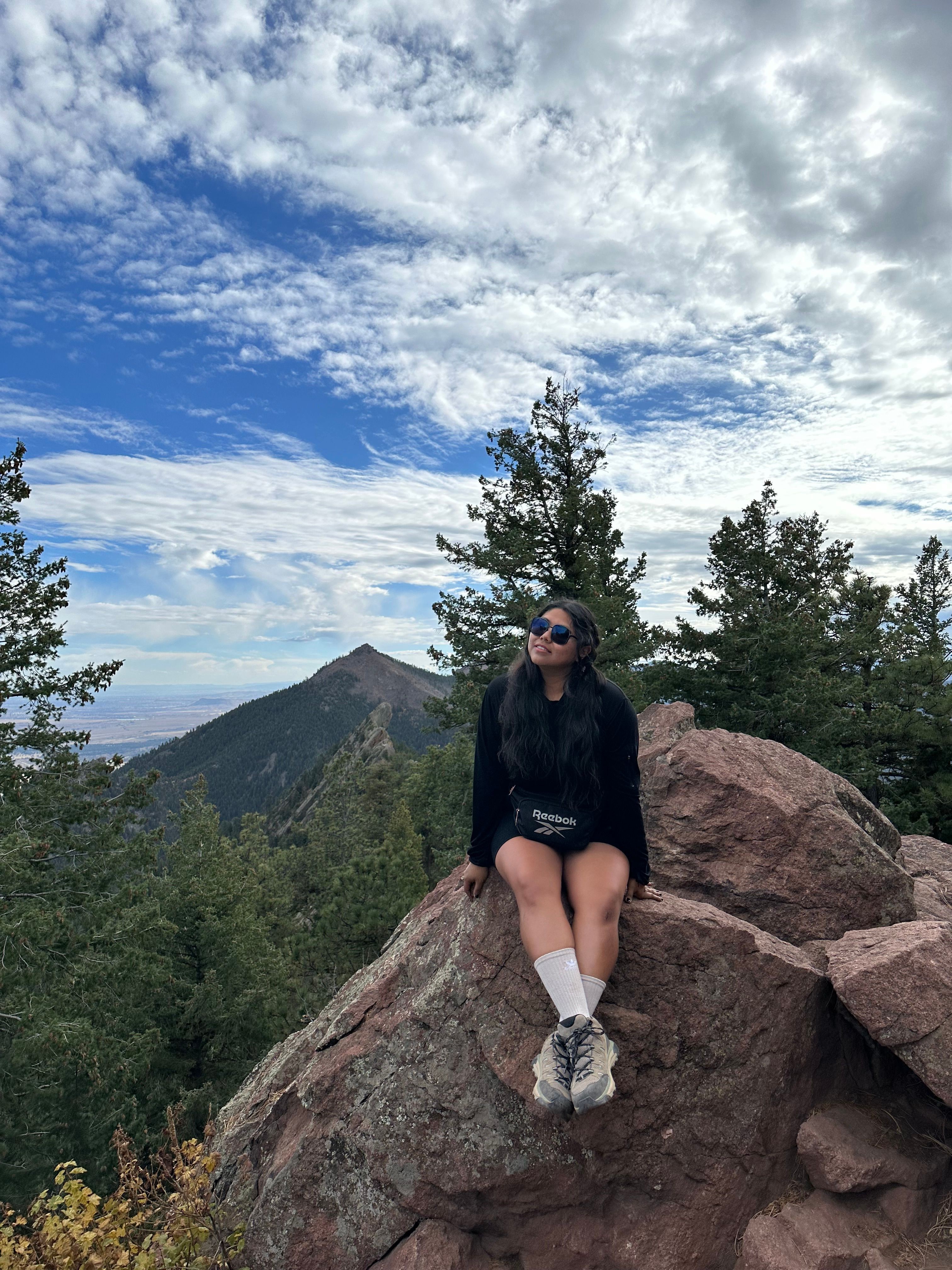 Person sitting on a large rock outdoors with trees and mountains in the background, under a partly cloudy sky.