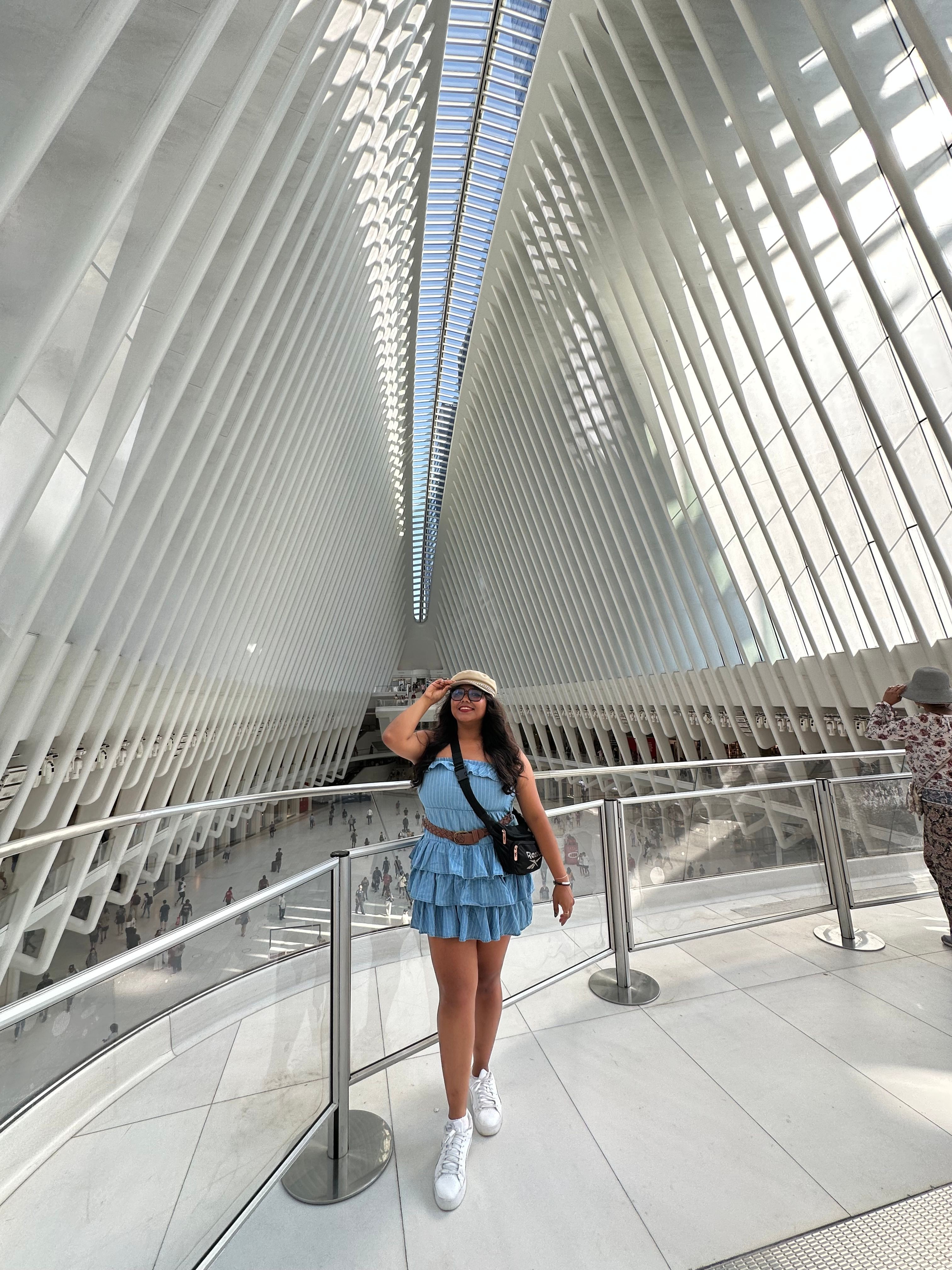 Young woman in a blue skirt and white sneakers standing on a glass walkway with metal railings, curved architecture, and vertical white slats.