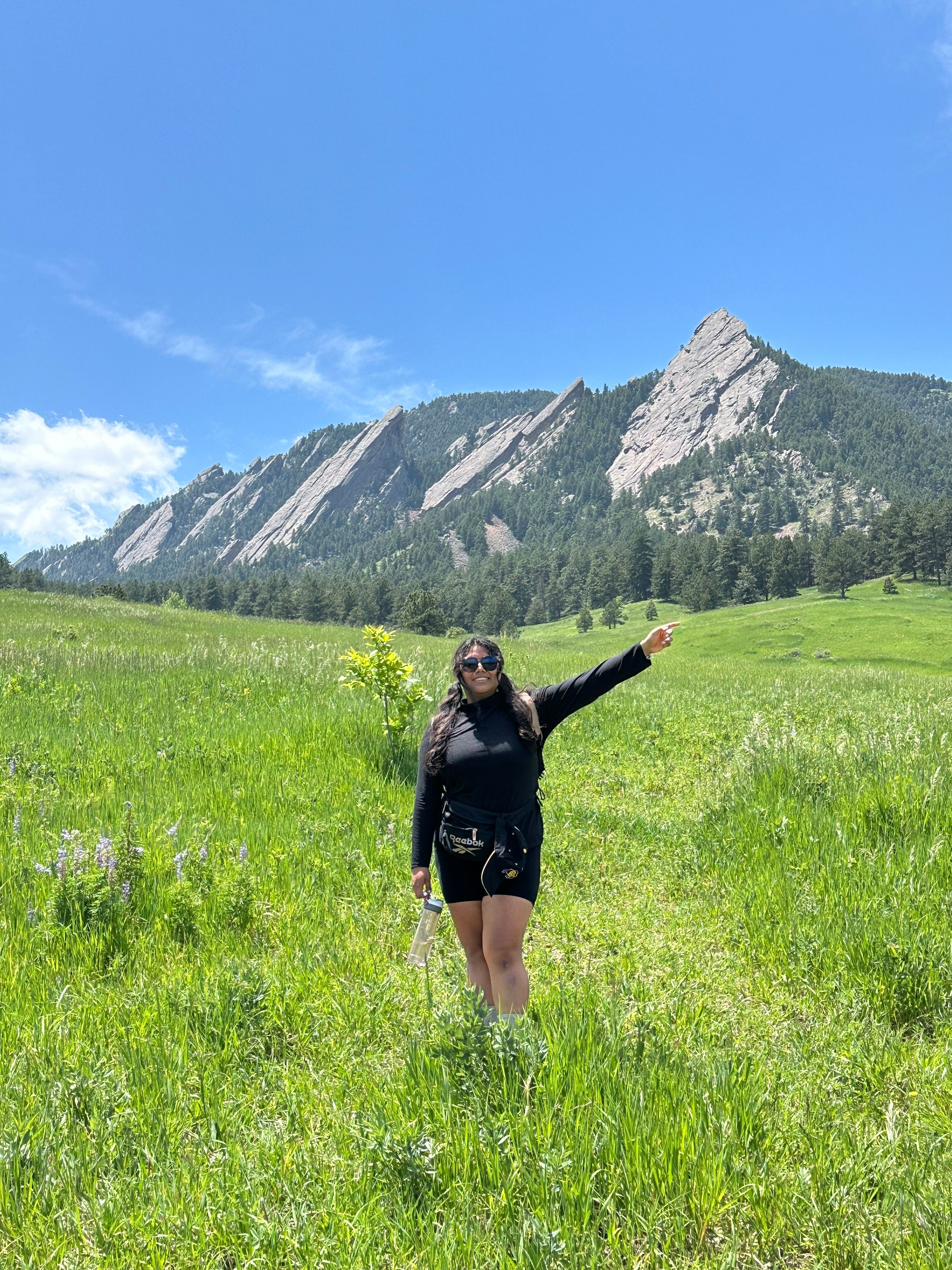 Person standing on grassy field with mountains and blue sky in background, raising one arm.