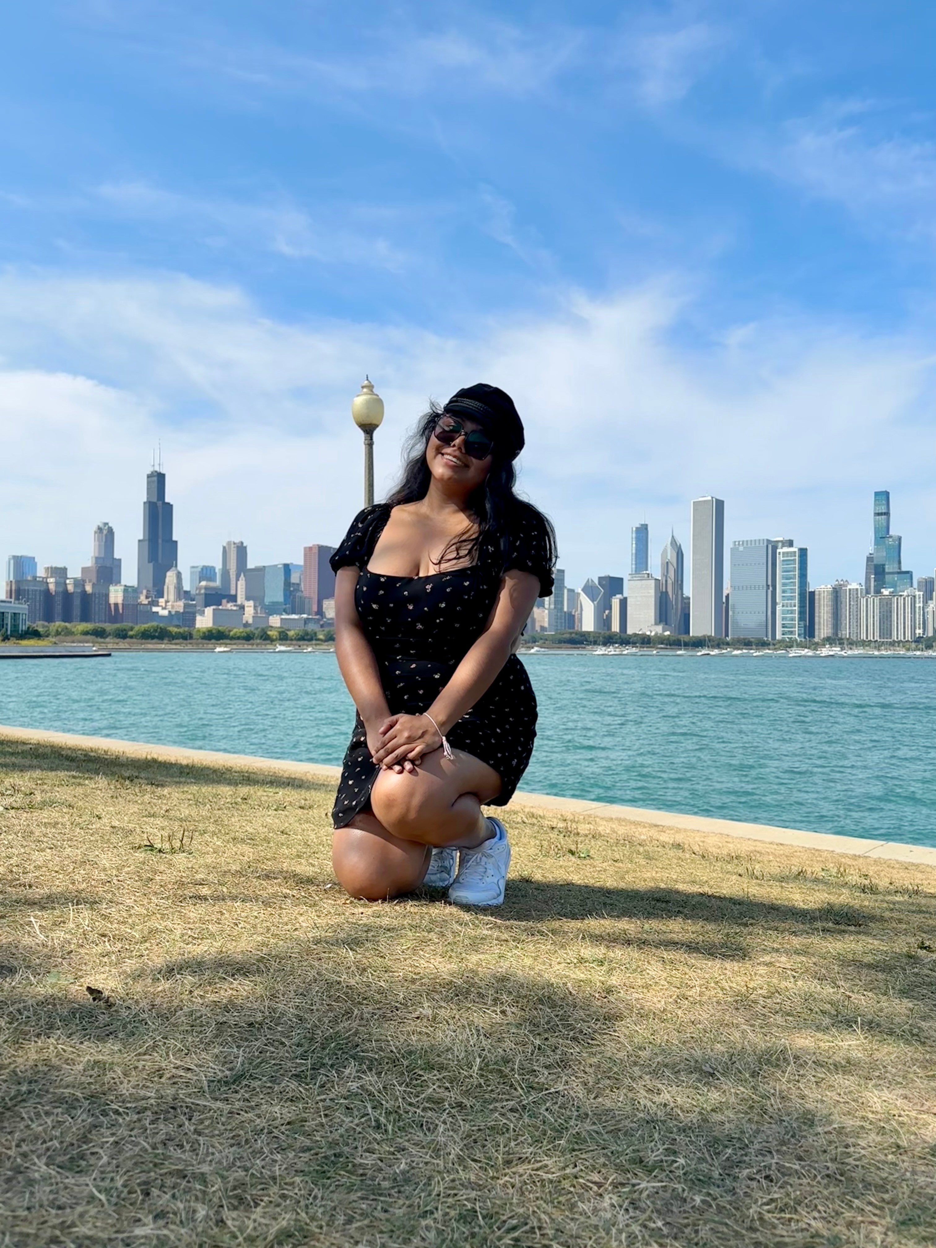 Woman kneeling on grass near water with city skyline in background, wearing black dress and sunglasses.