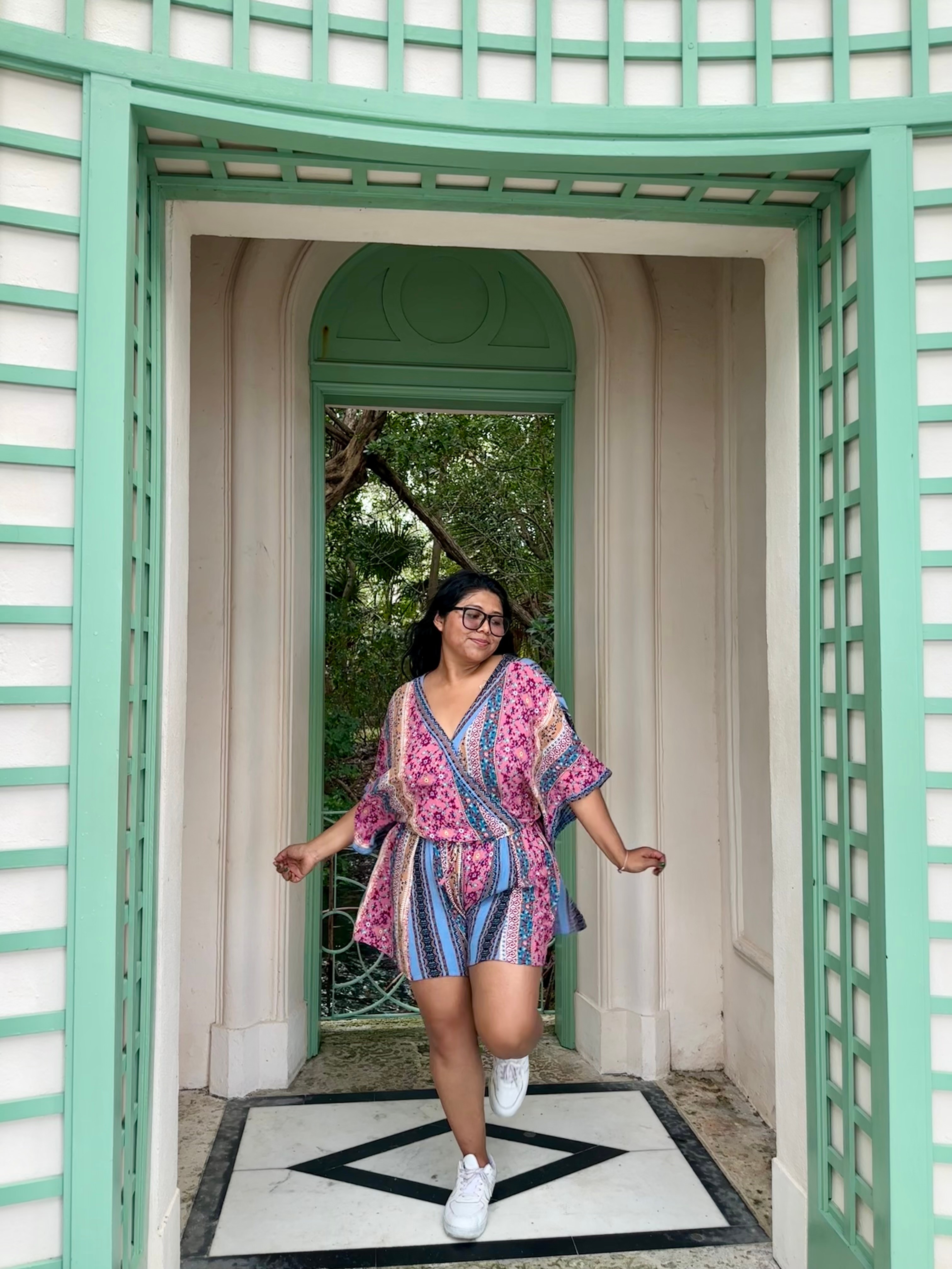 Woman in colorful dress standing in an arched doorway with green framing, greenery visible behind her.