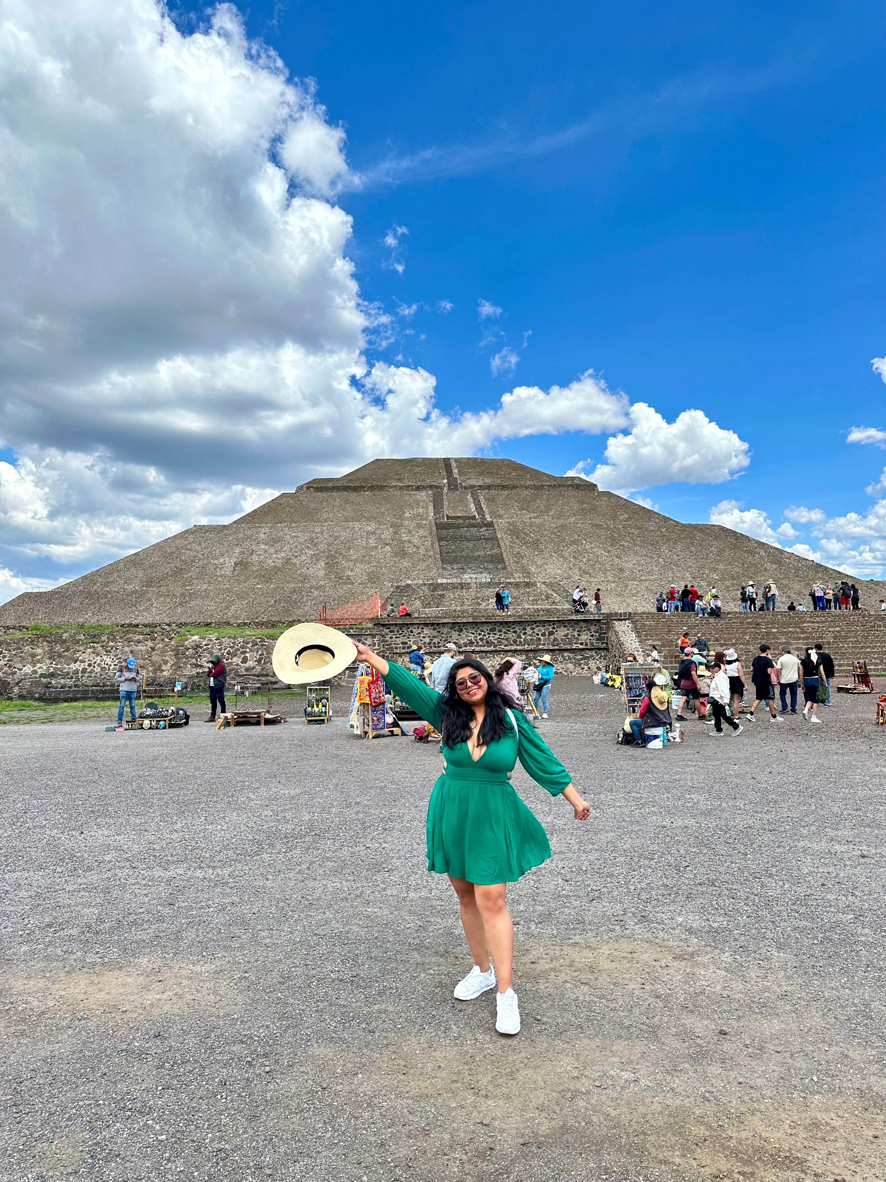Young woman in green dress and white shoes standing in front of a large pyramid, with people and a blue sky with clouds.