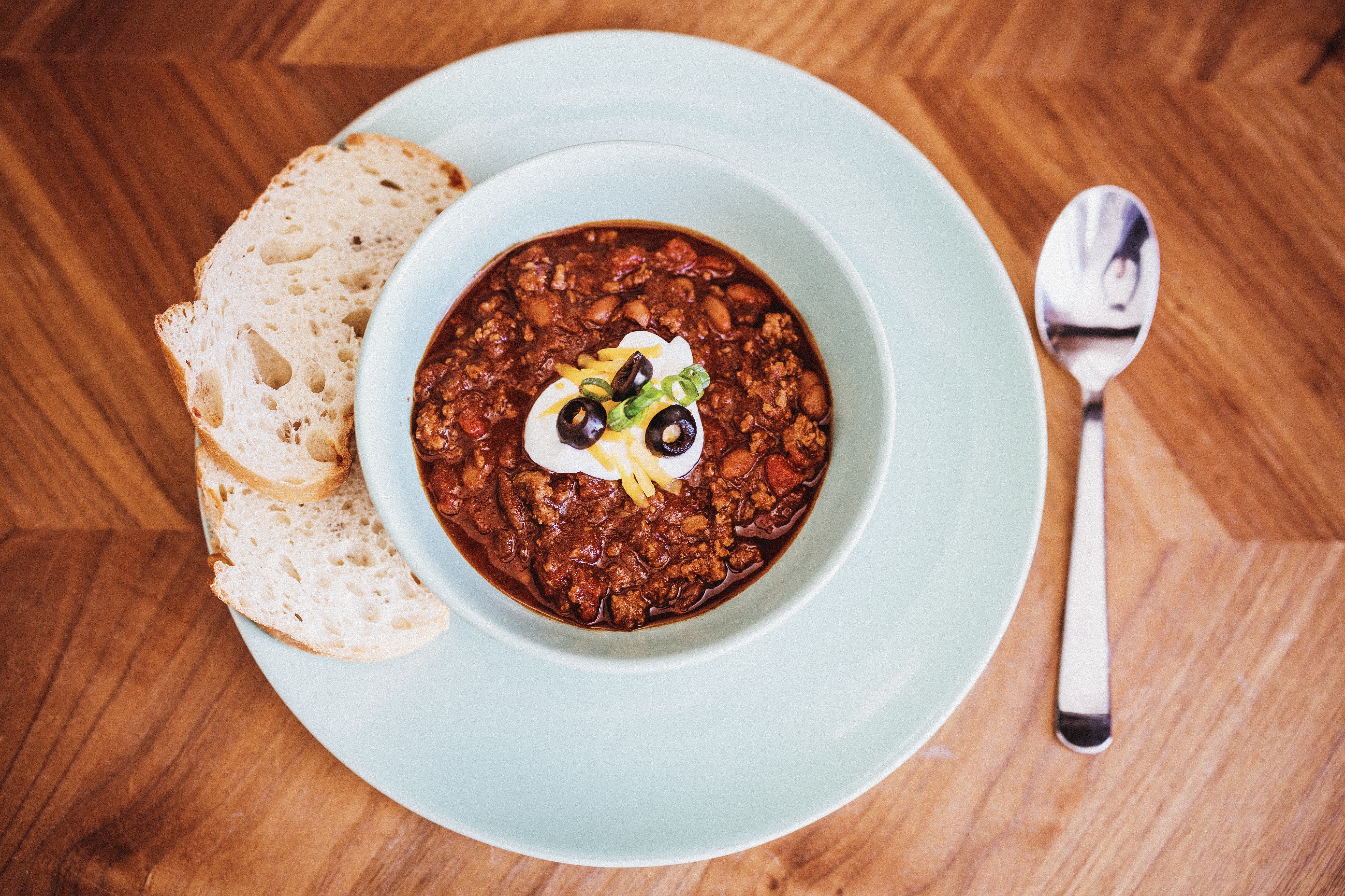 Bowl of chili garnished with sour cream and black olives, served with bread slices on a white plate, spoon on the right.