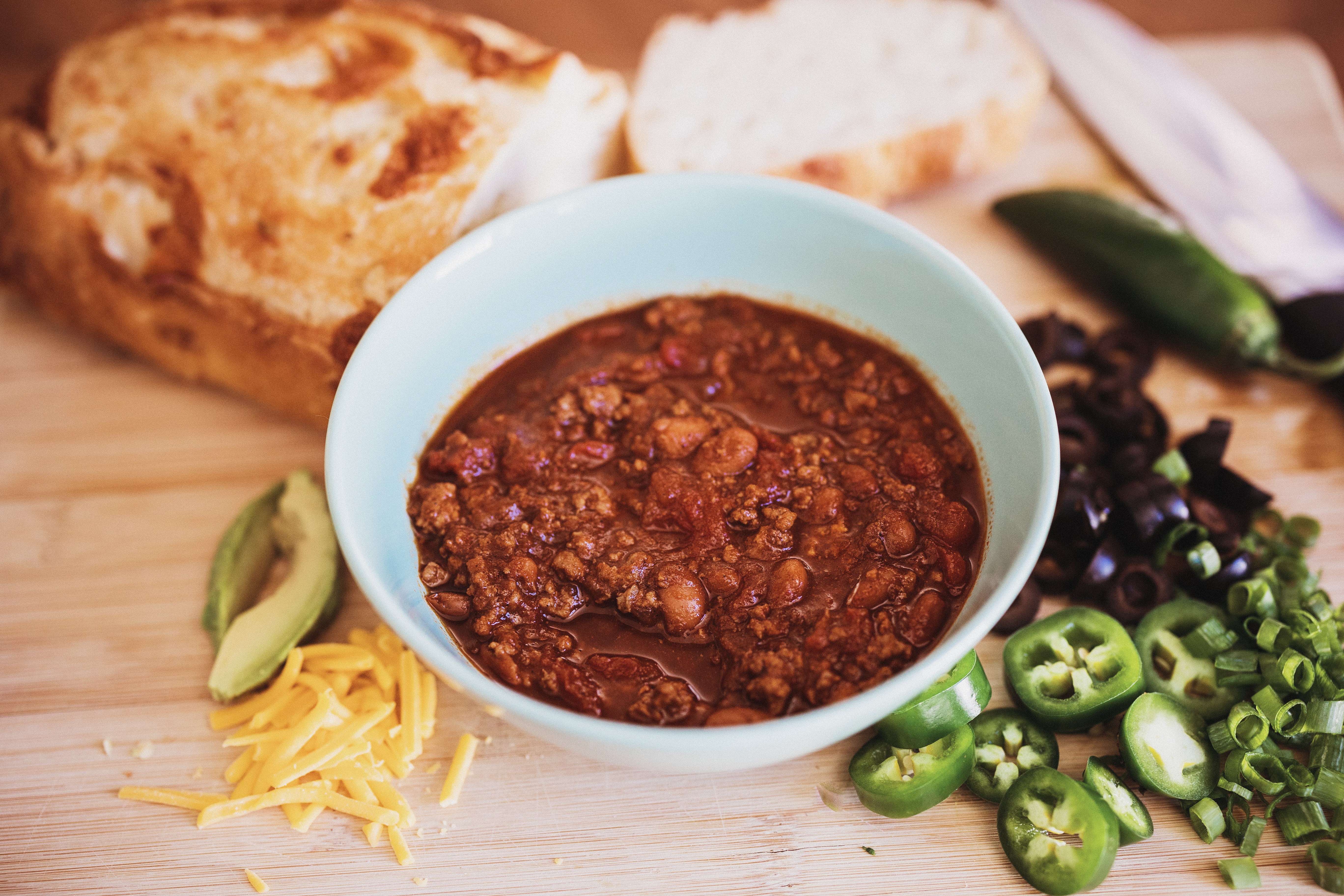 Bowl of chili with bread, avocado, shredded cheese, and sliced green peppers on a wooden surface.