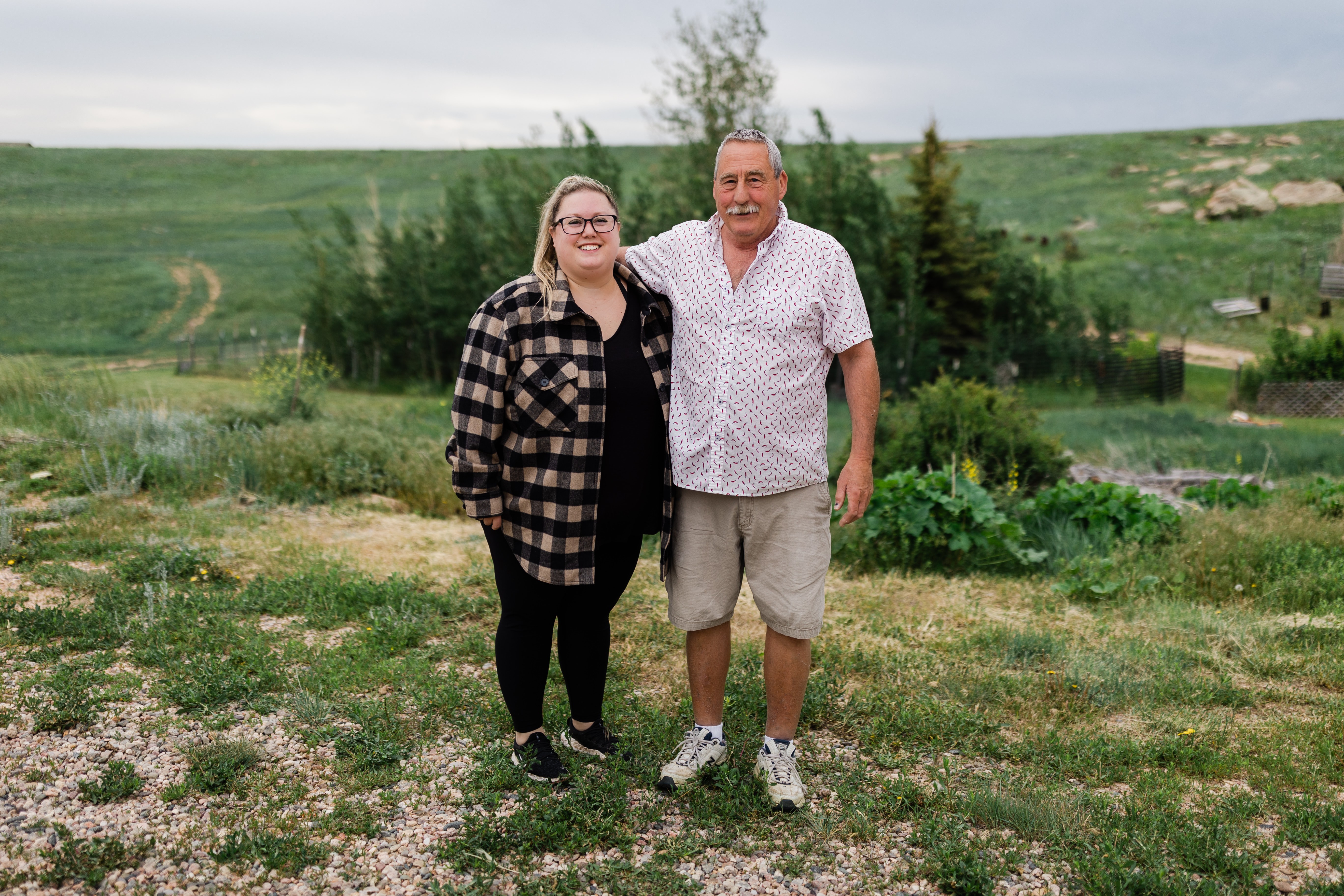 Two people standing outdoors on grassy terrain with trees and hills in the background.