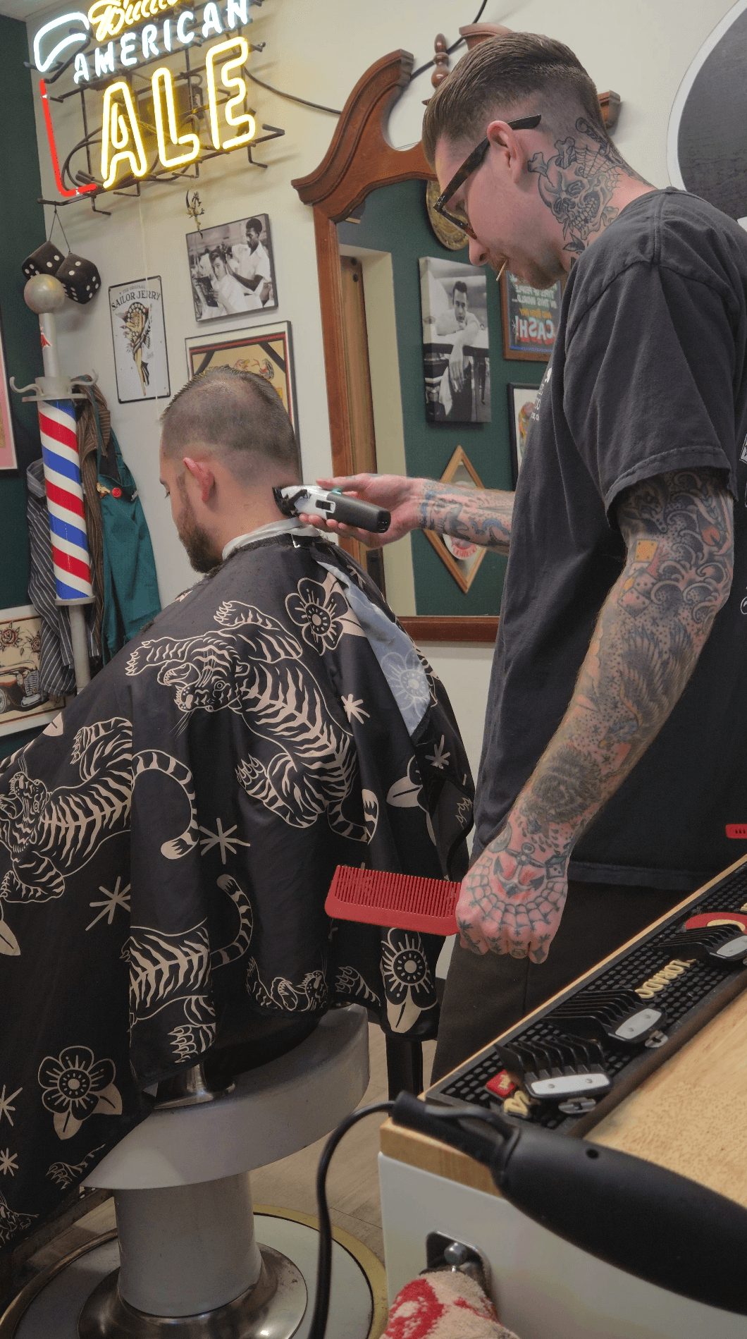 Barber trims customer's hair in a barbershop with vintage decor and a neon sign reading 'American Ale'.