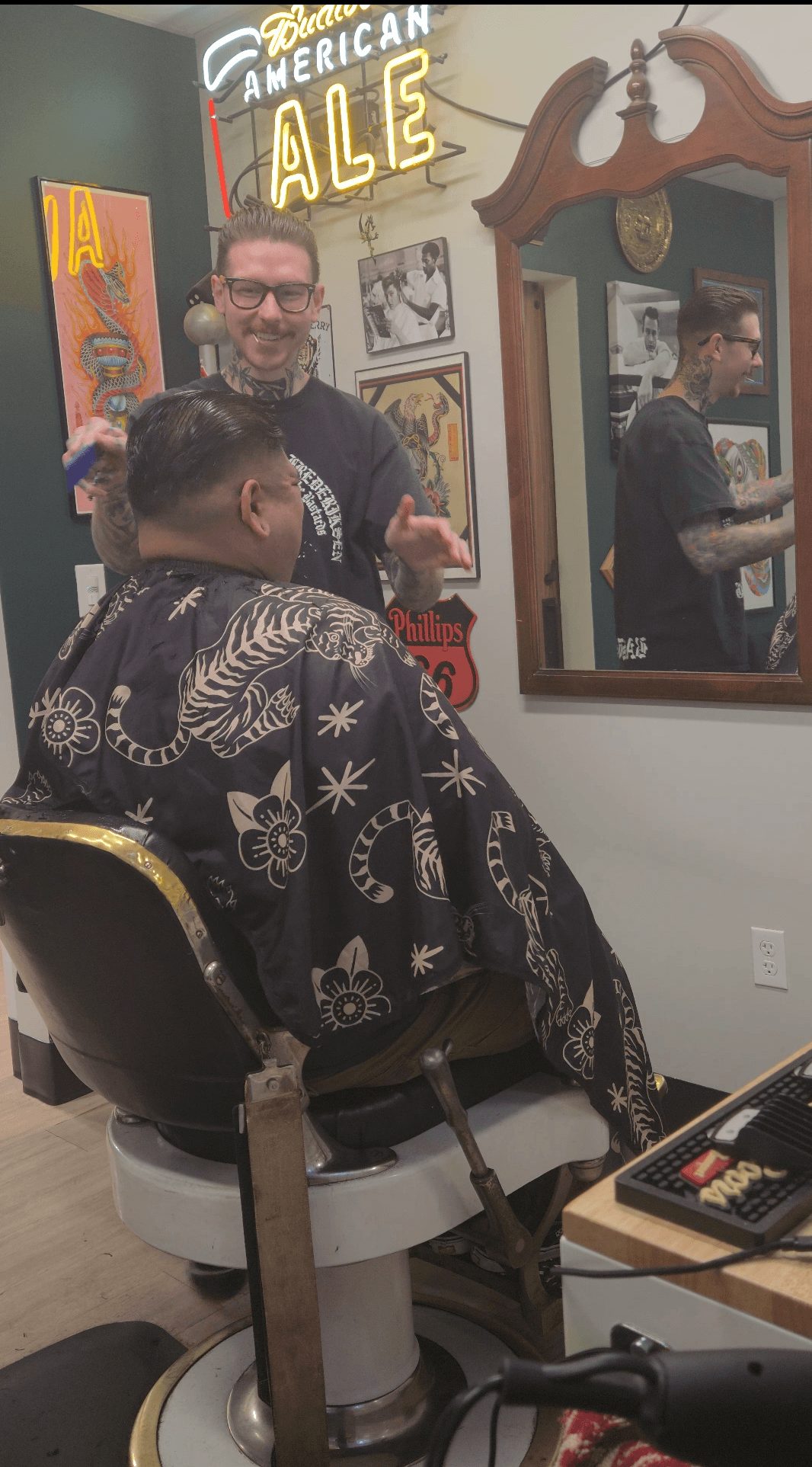 Person getting a haircut in a salon, with a barber smiling and holding scissors, mirror reflecting the scene, and salon decor visible.
