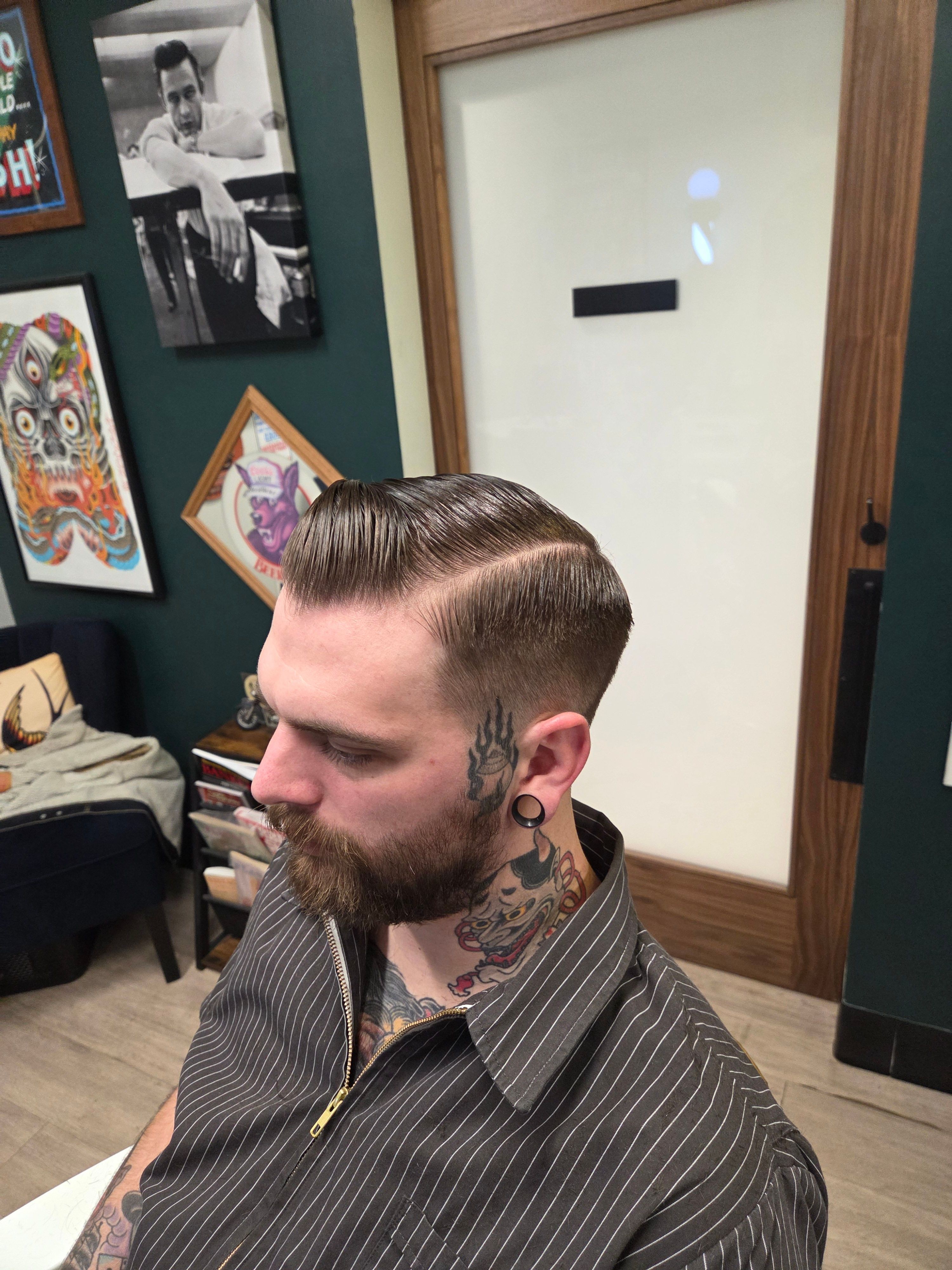 Side view of a man with a slicked-back hairstyle and beard, wearing a striped shirt, in a room with framed pictures on the wall.
