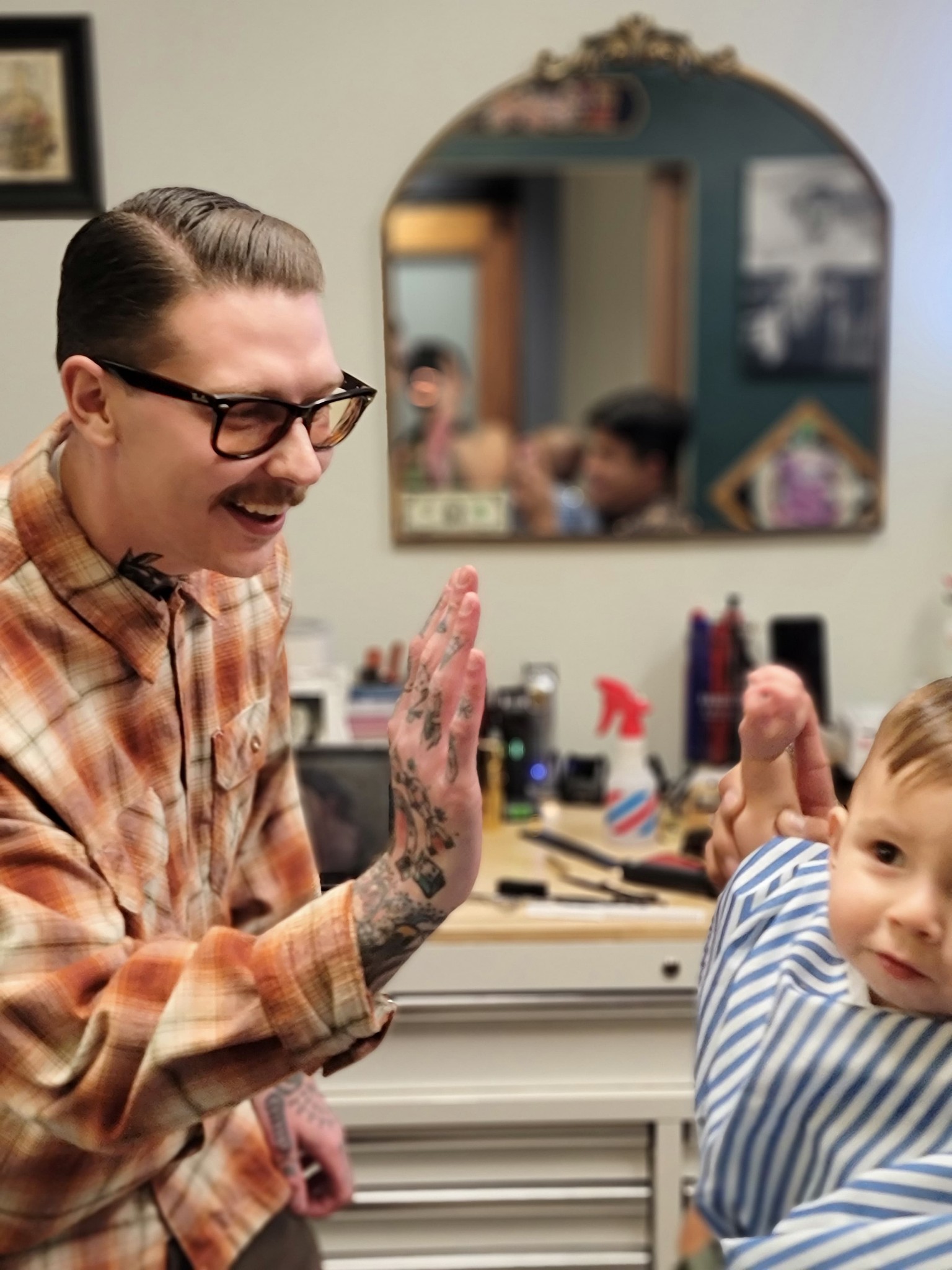 Person with glasses and tattoos smiling and giving a high five to a young child in a barbershop, mirror and tools in background.