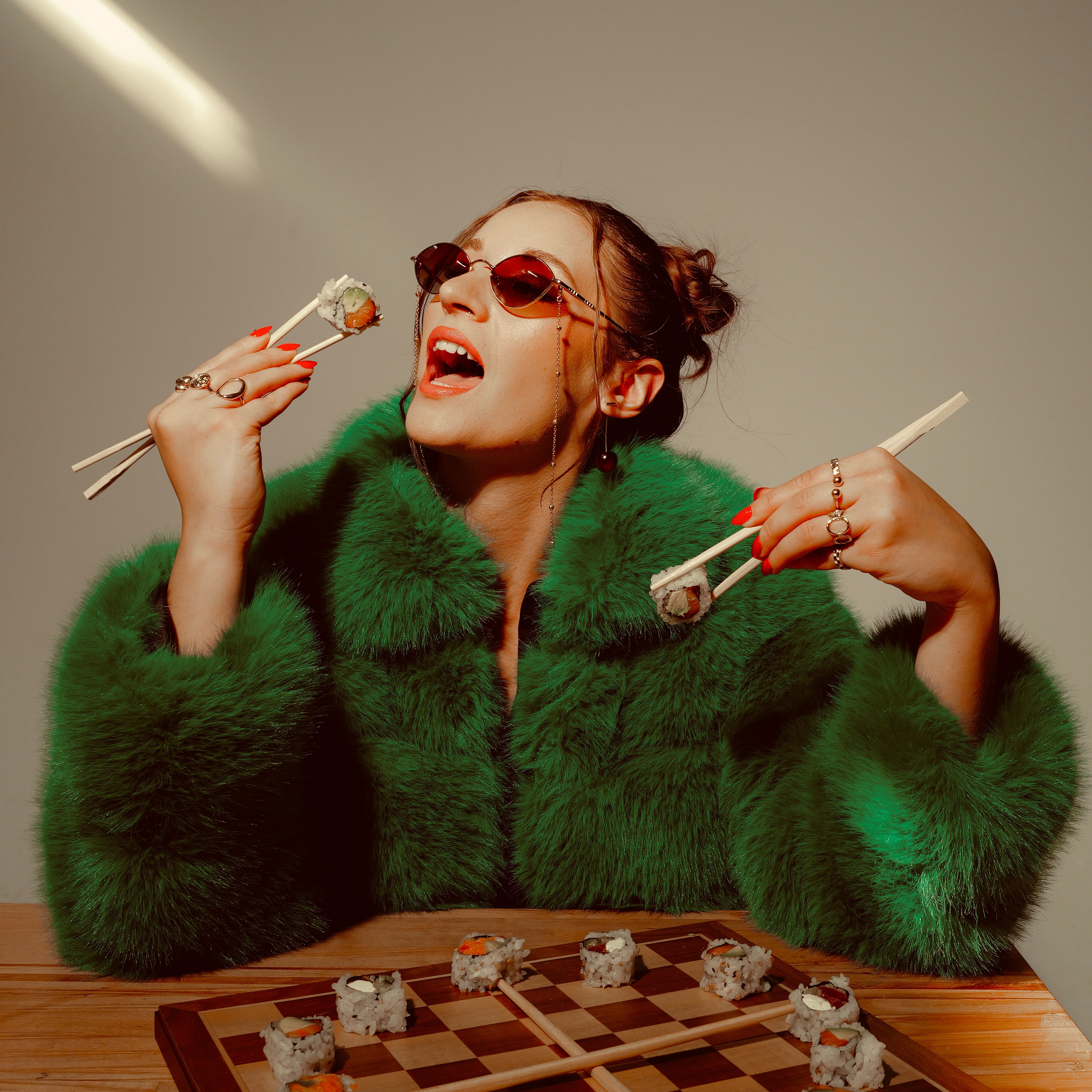 Woman with sunglasses and green fur coat holding sushi and chopsticks, smiling, sitting at a table.