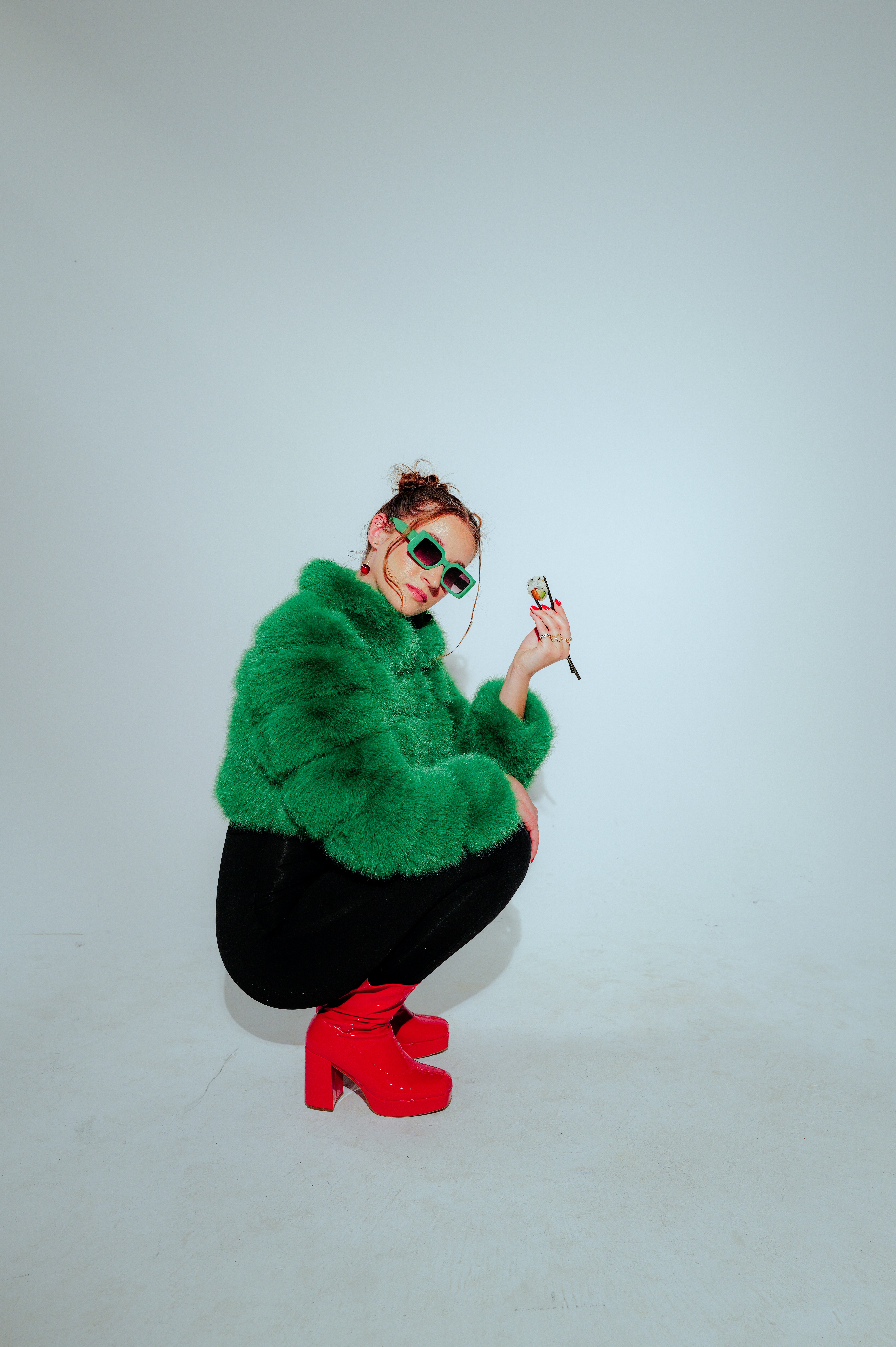 Person crouching in green fur coat, red boots, holding a phone, wearing sunglasses and makeup, against a plain background.