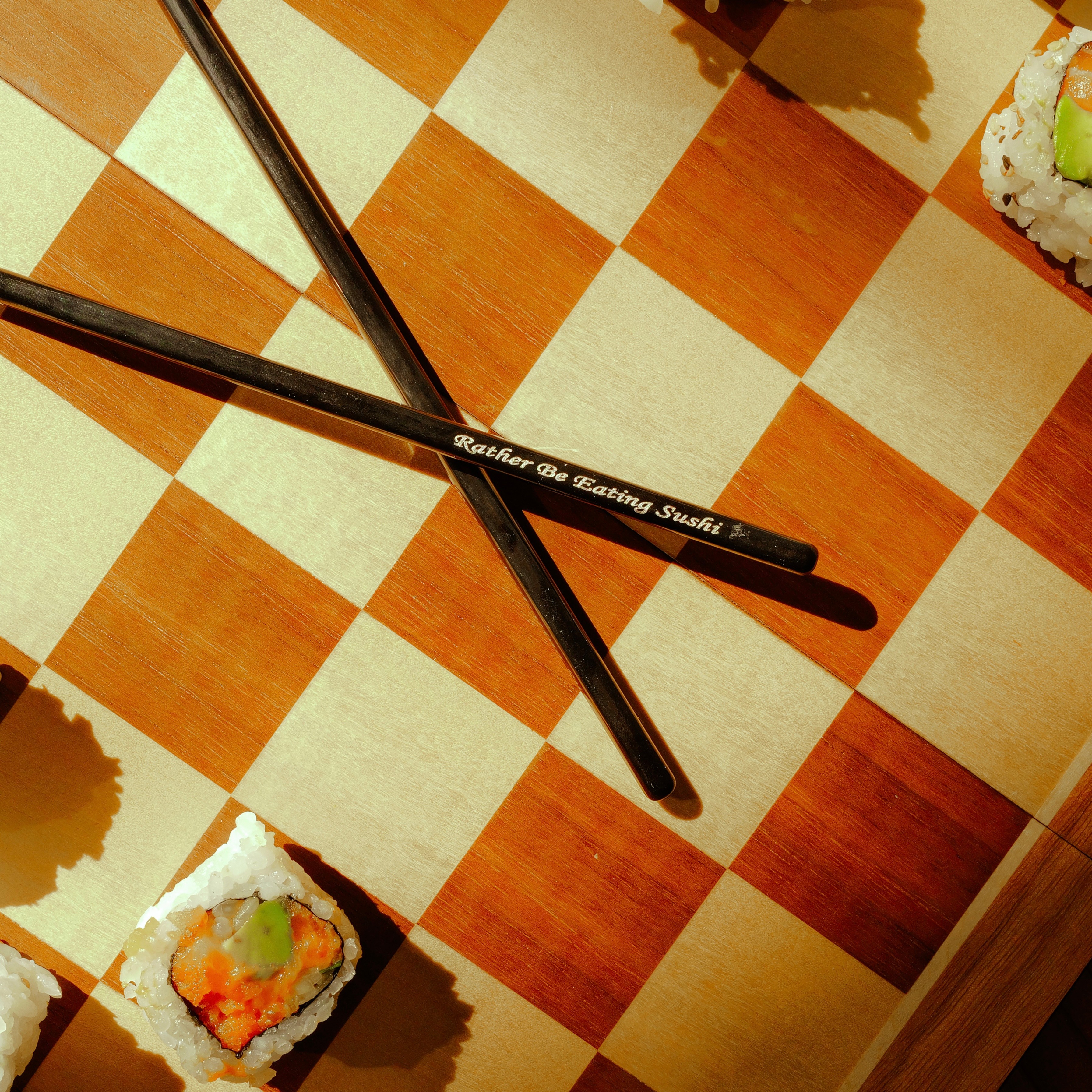 Two black chopsticks crossed on a checkered orange and white table, with sushi rolls partially visible in the corners.