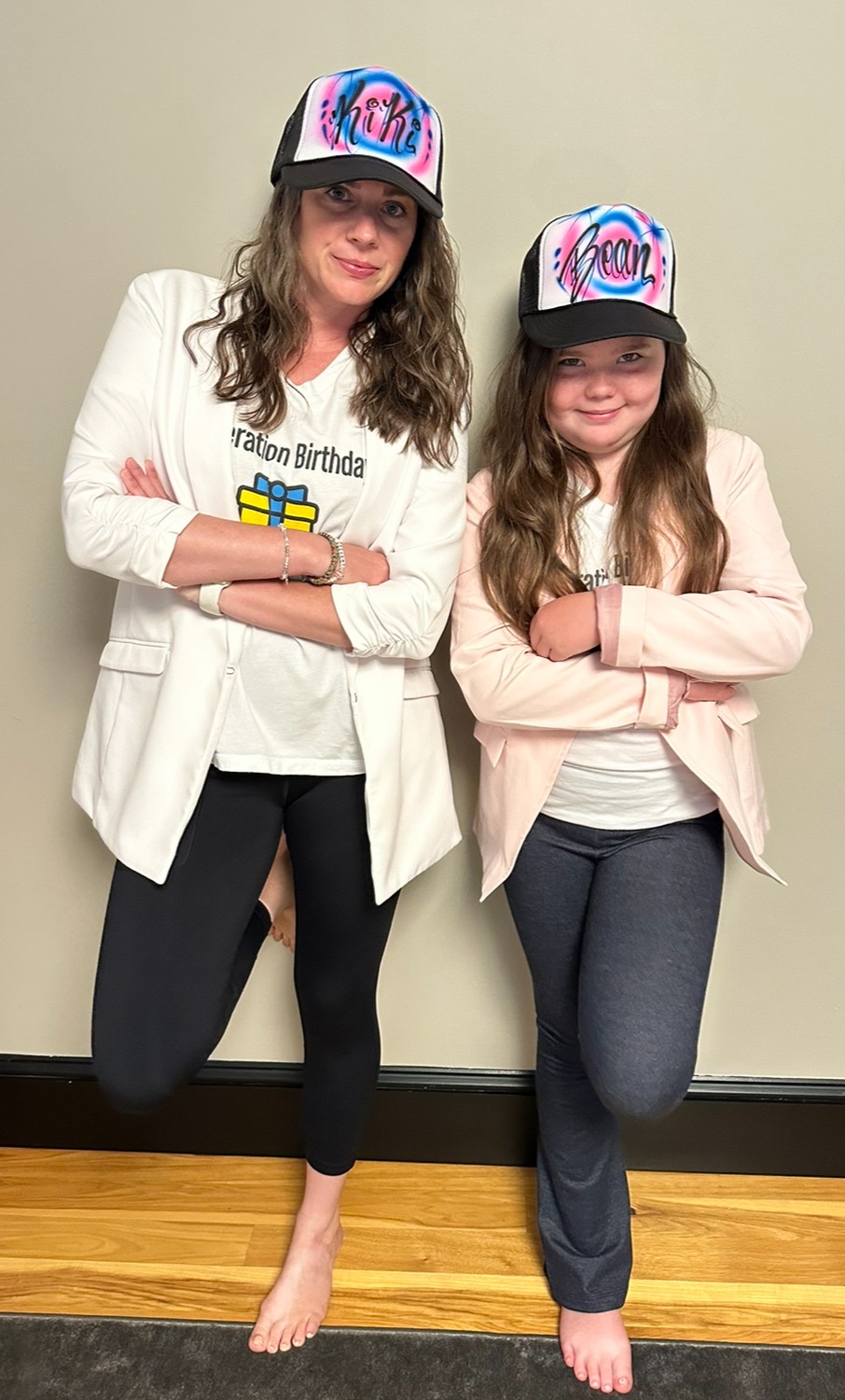 Two girls stand side by side, wearing matching birthday hats and casual clothing, smiling with arms crossed, against a plain wall.