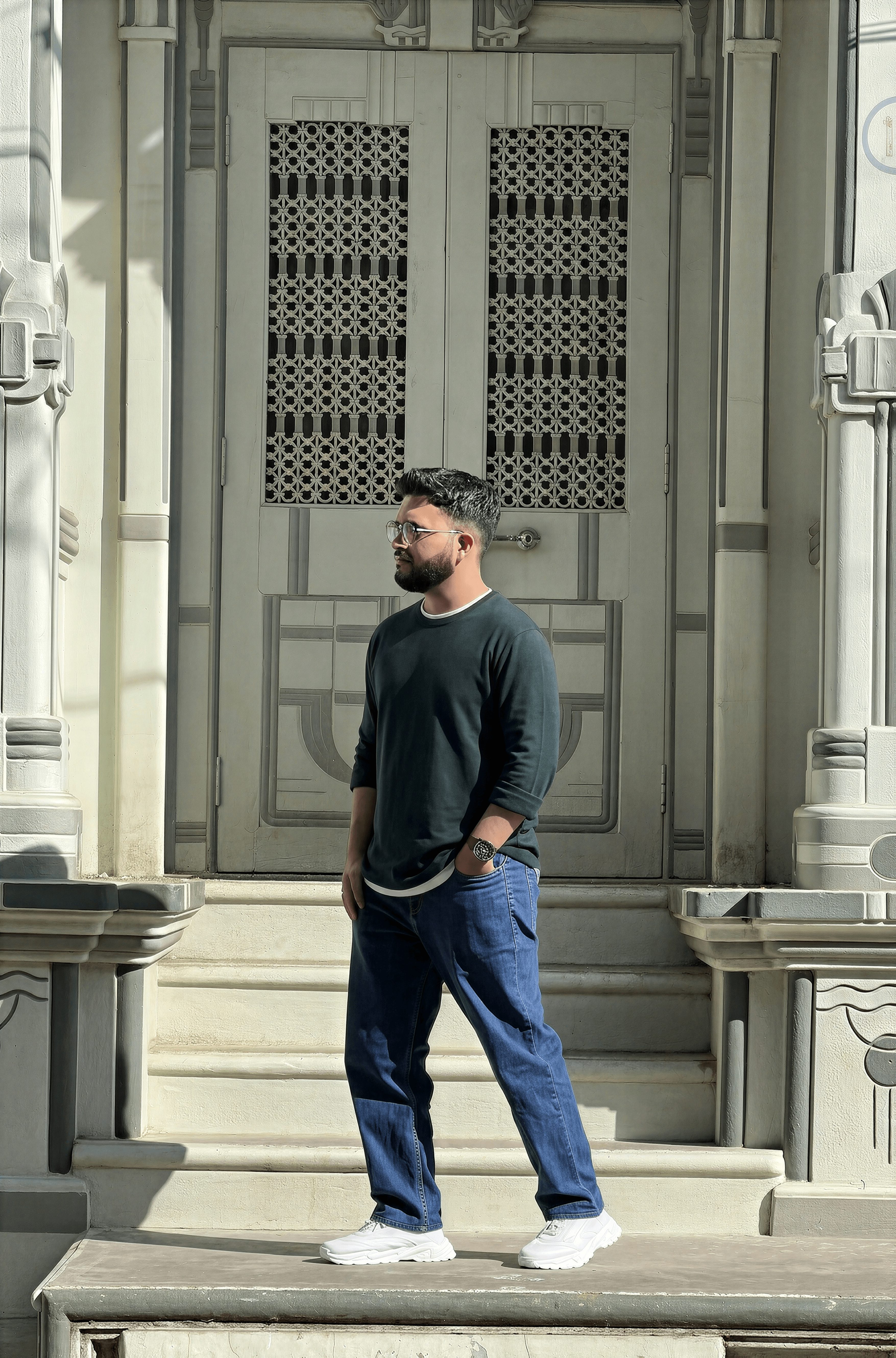 Man with beard in dark sweater and jeans stands on steps in front of a large ornate door.
