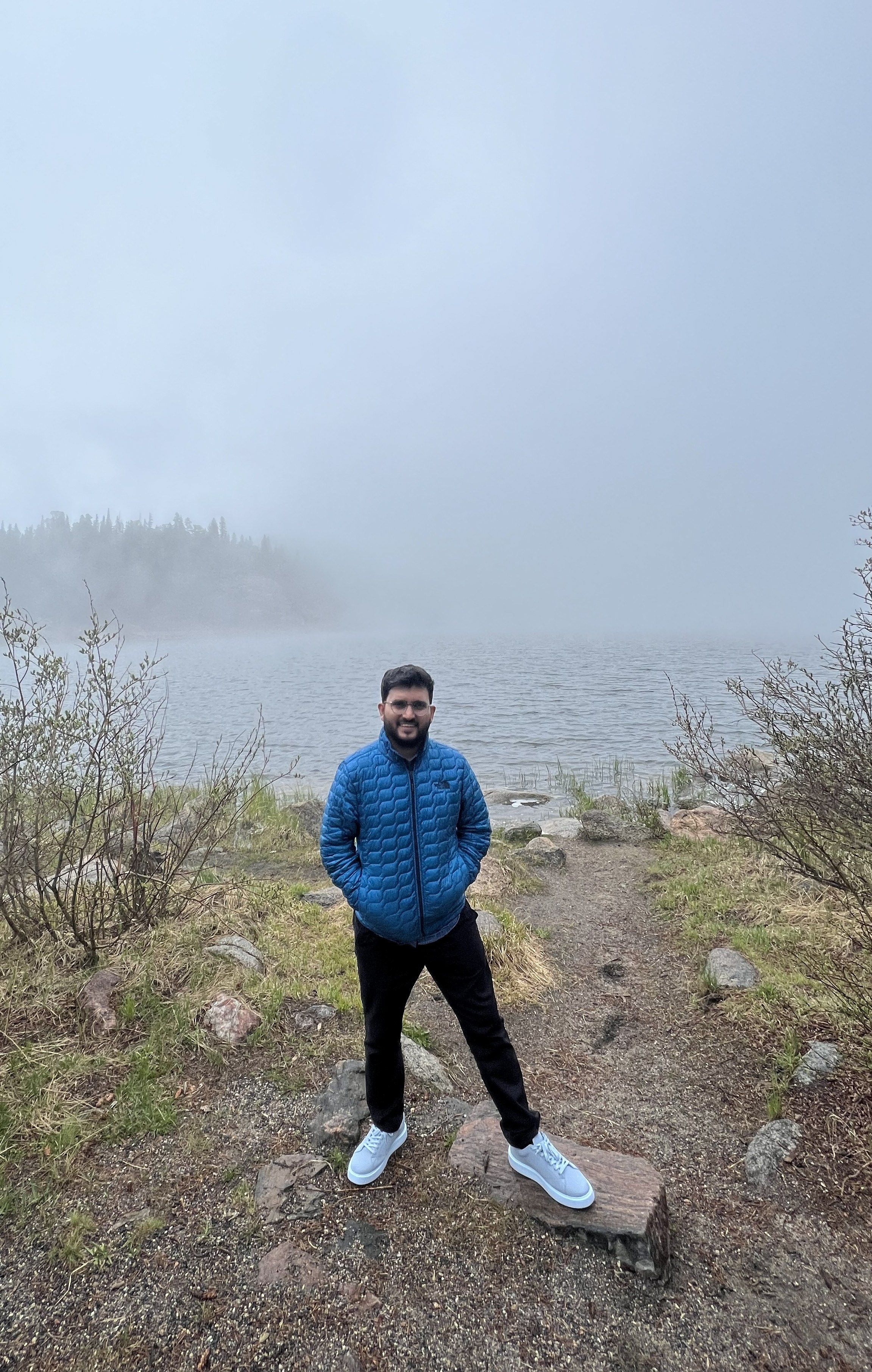 Person standing outdoors near a lake with foggy sky, wearing a blue jacket, black pants, and white shoes.