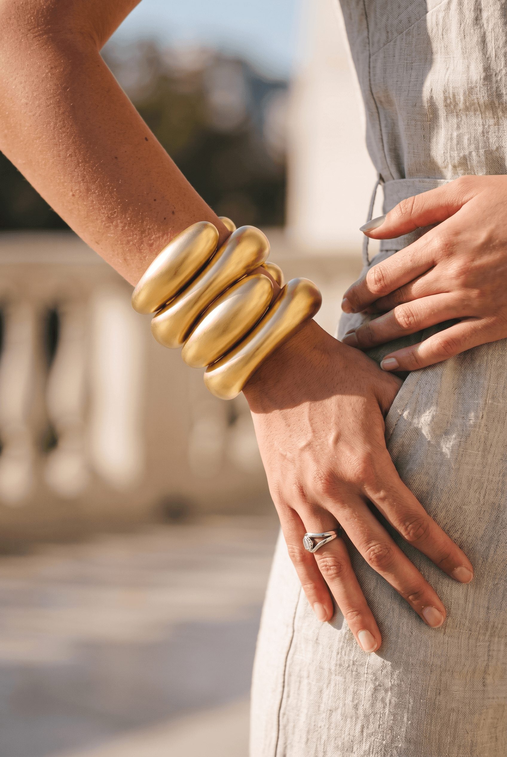 Close-up of a person wearing large gold bangles and a ring, with their hand resting on their hip, outdoors.