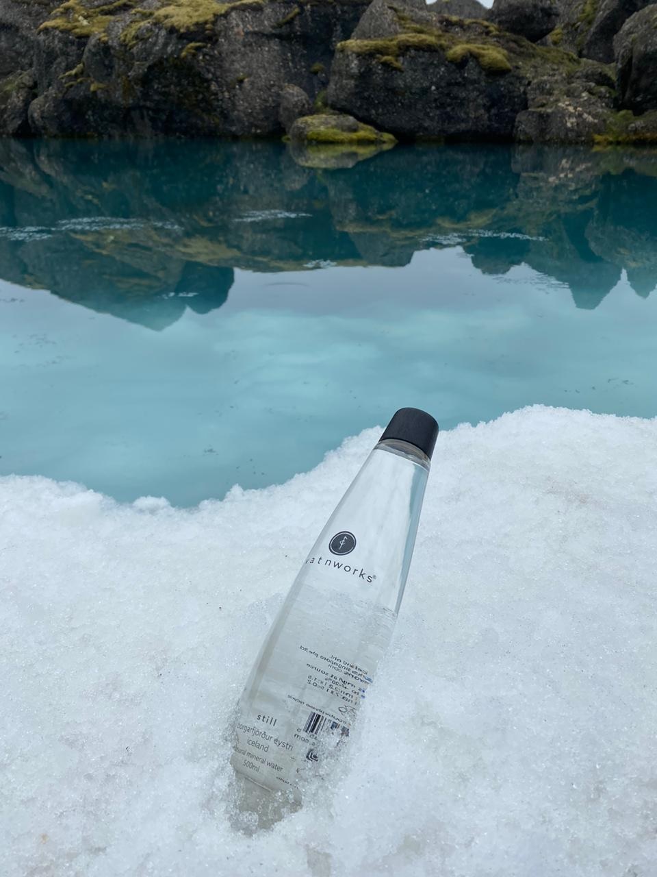 A clear plastic water bottle with a black cap lies on snow near a body of water with rocks and moss in the background.