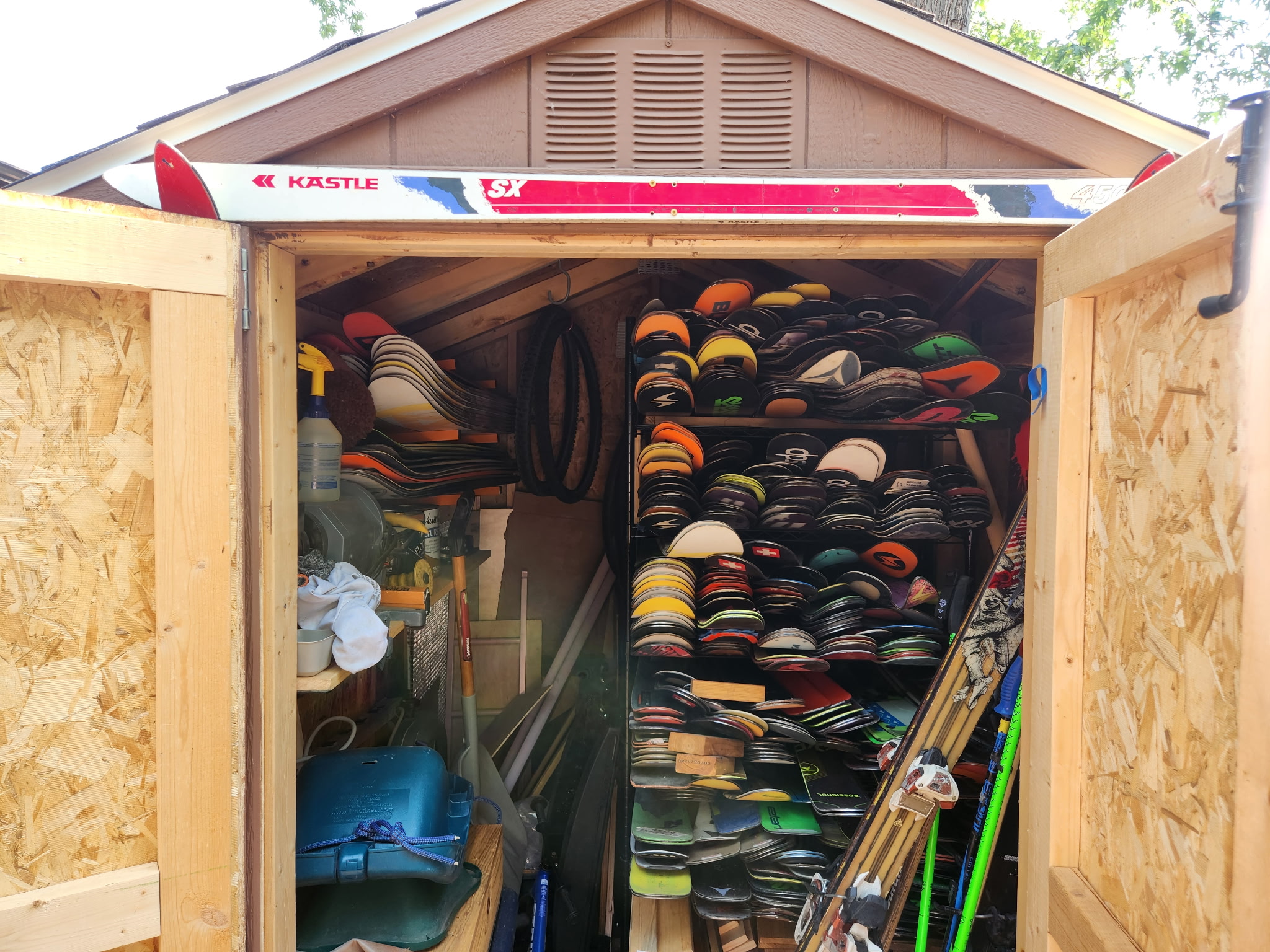 Open shed door showing shelves filled with helmets and equipment, with a ladder leaning inside, and a building in the background.