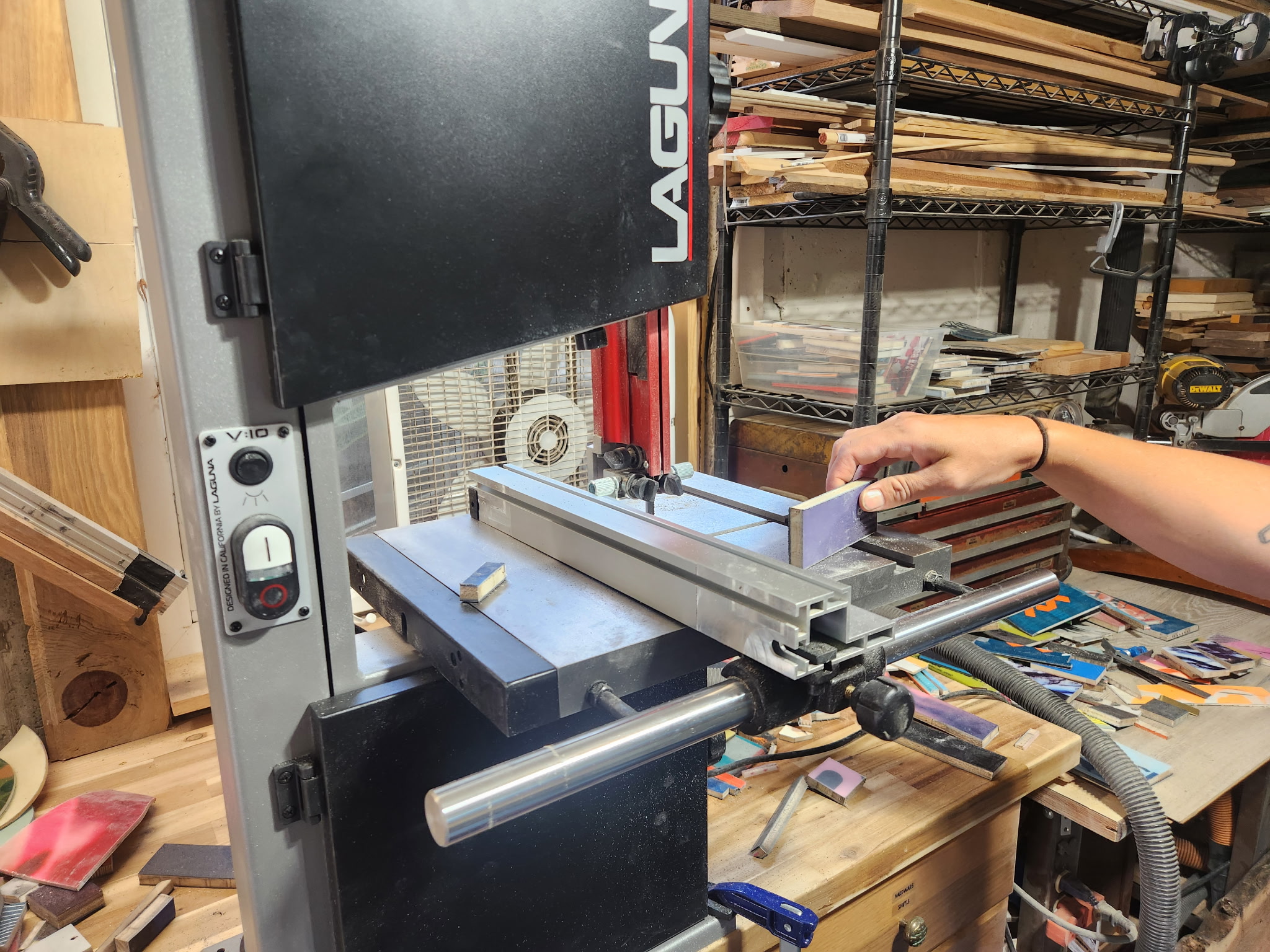 Person's hand operating a metal cutting machine in a workshop with tools and materials on shelves and workbench.