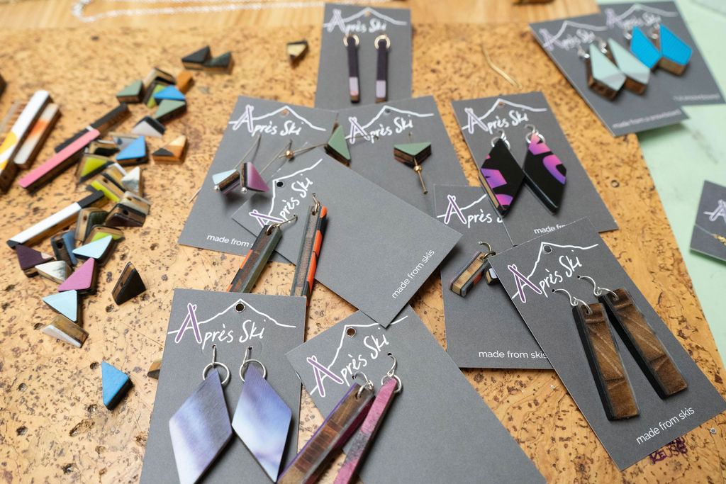 Multiple jewelry earrings on black cards, some scattered on a wooden surface, with colorful geometric earrings and packaging visible.