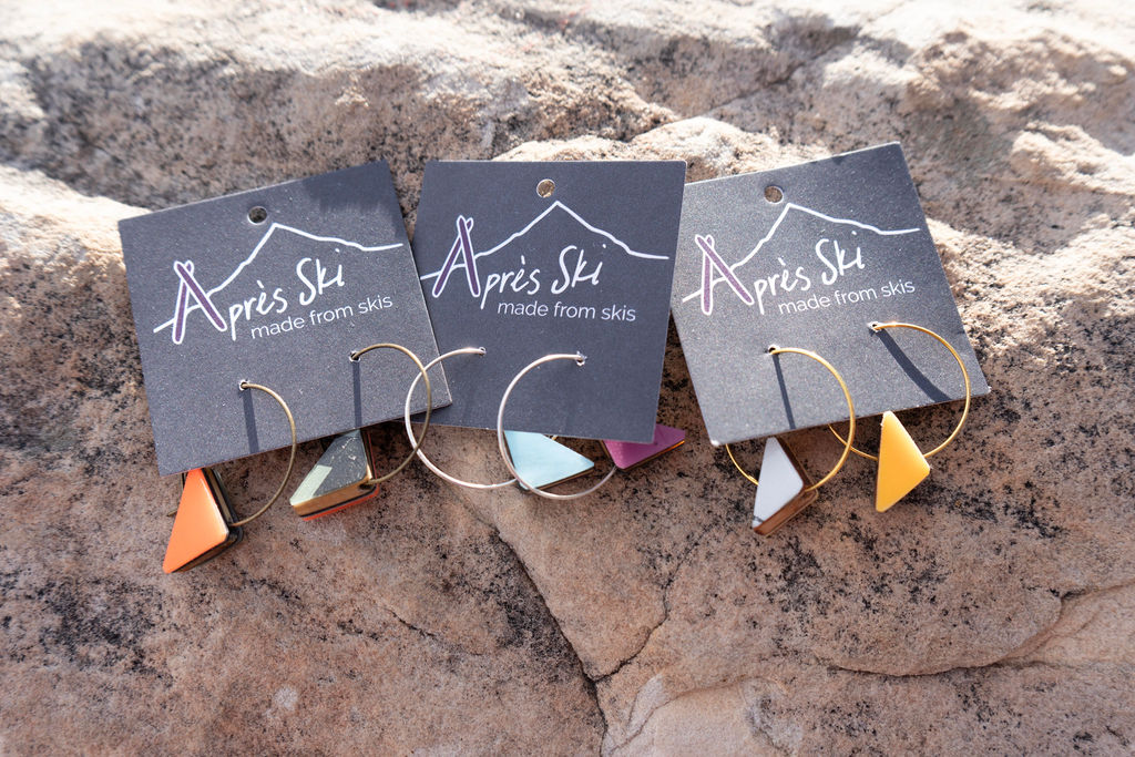 Three pairs of earrings with colorful triangle pendants on a textured surface, each on a black card with white text.