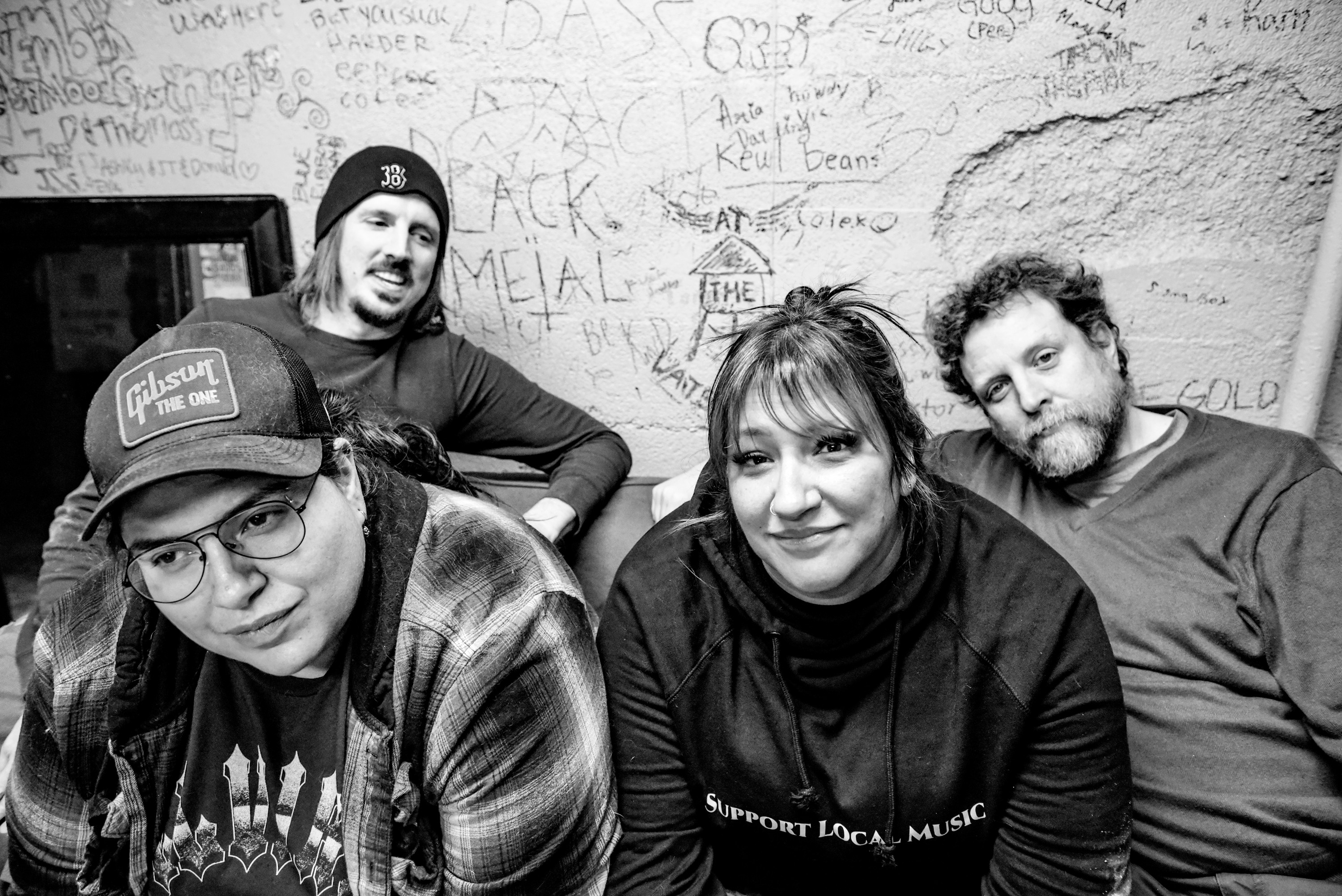 Four people sitting against a wall with graffiti, smiling and looking at the camera.