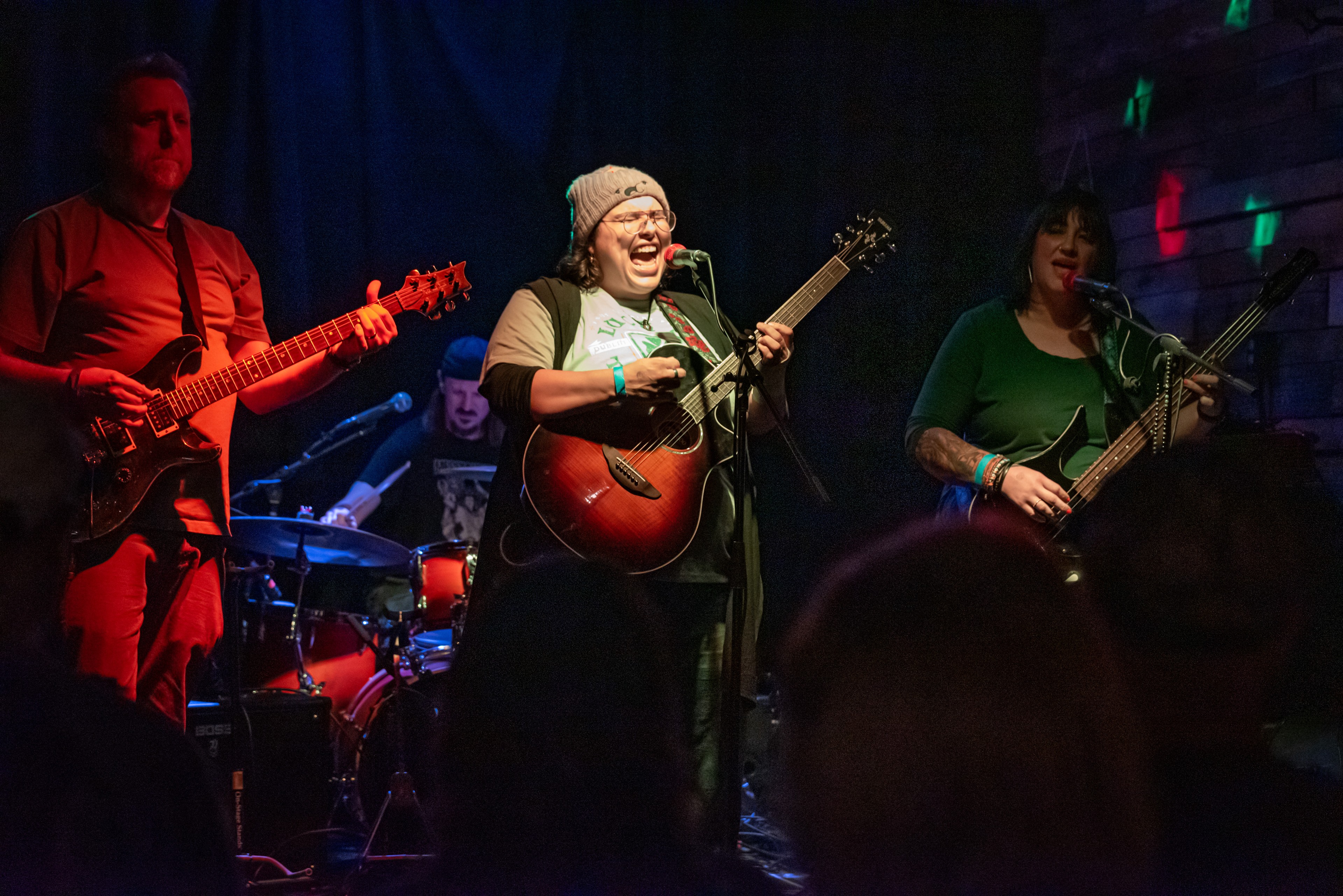 Musicians perform on stage with colorful lighting, including a woman singing and playing guitar, and others playing instruments.