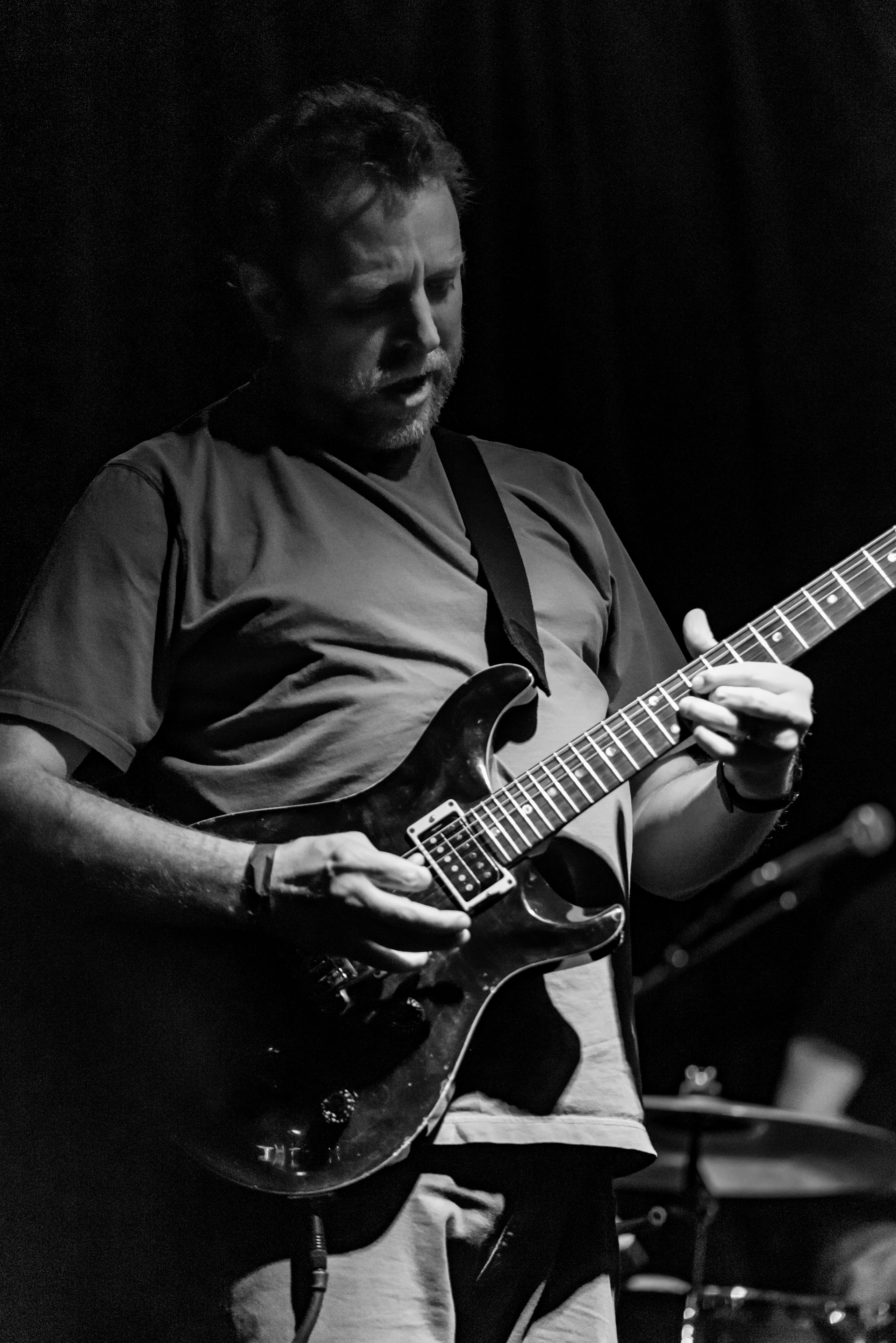 Man playing electric guitar on stage with dark background, focused expression, wearing a t-shirt, in black and white.