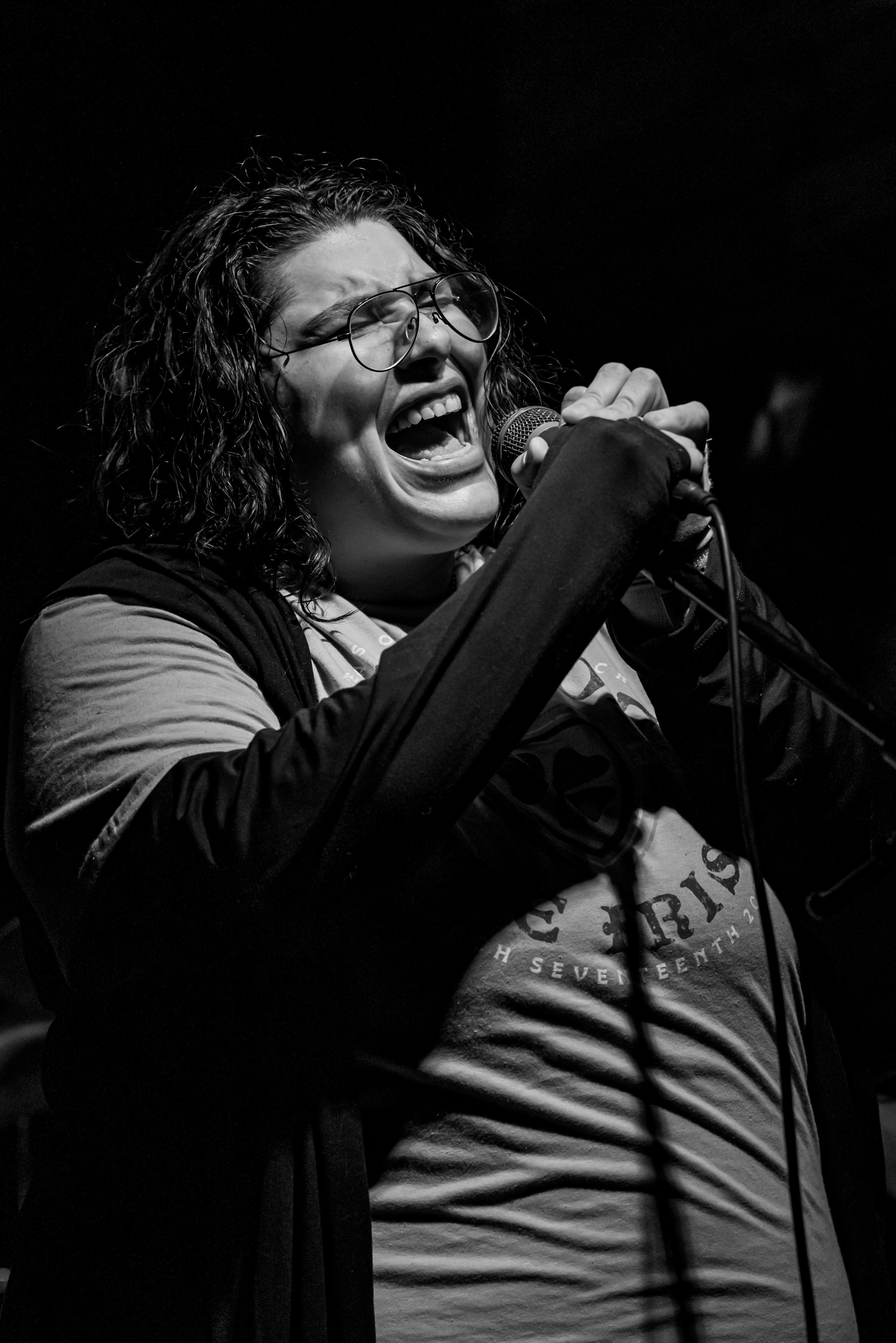 Person singing passionately into a microphone with curly hair and glasses, in black and white.