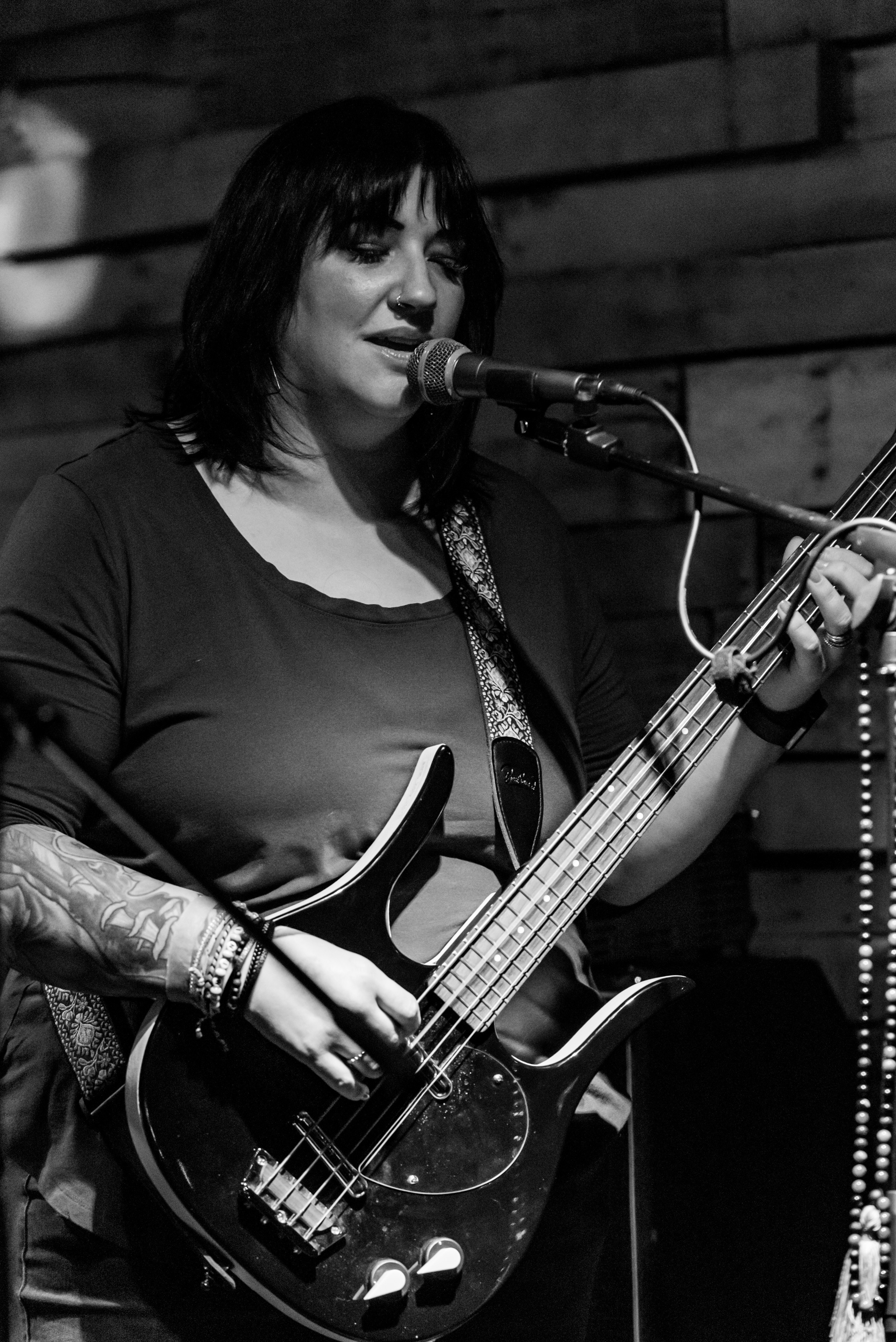 Woman with dark hair playing bass guitar and singing into microphone in a wooden room, black and white image.