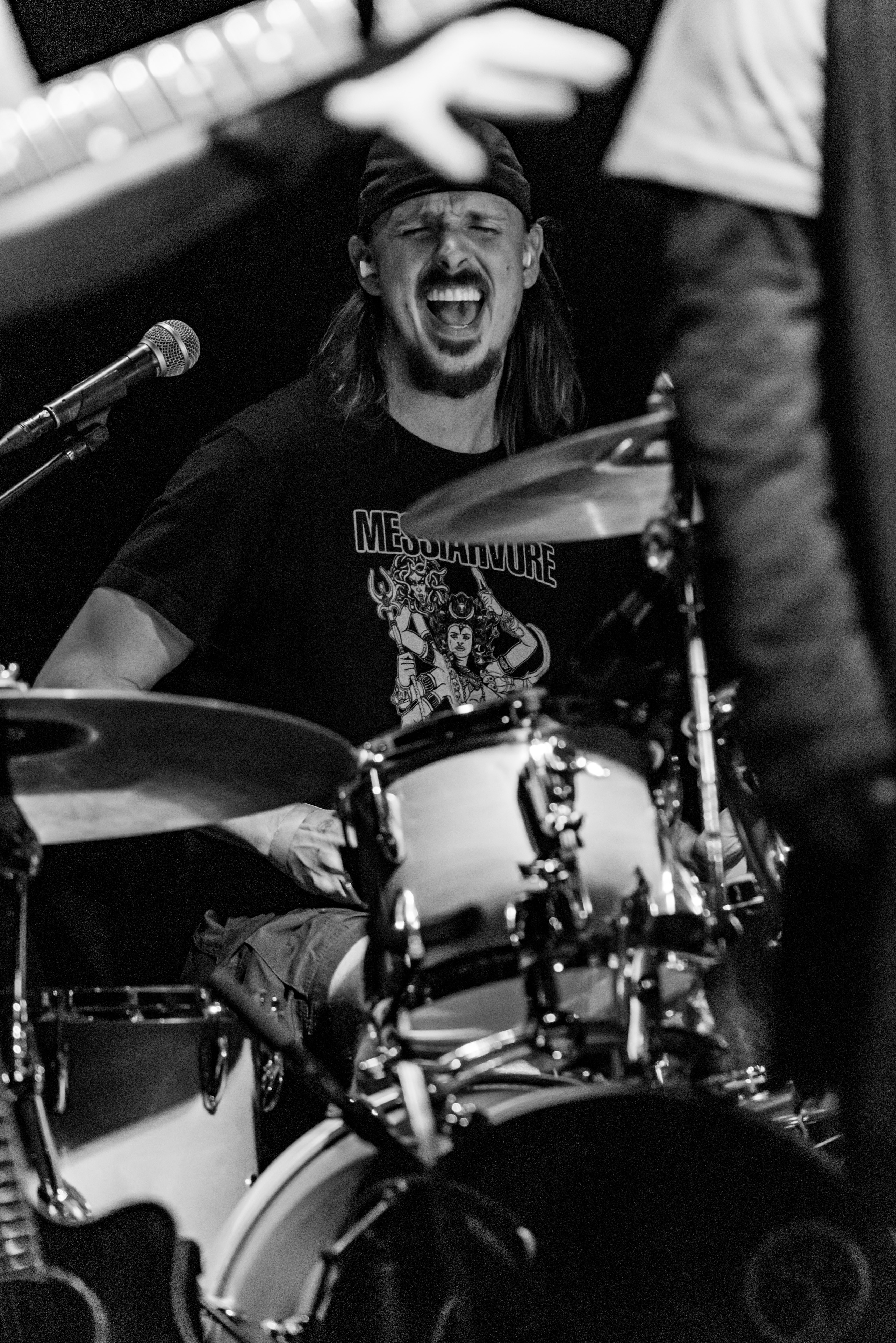 Person playing drums, smiling, with a microphone nearby, in a black and white setting.