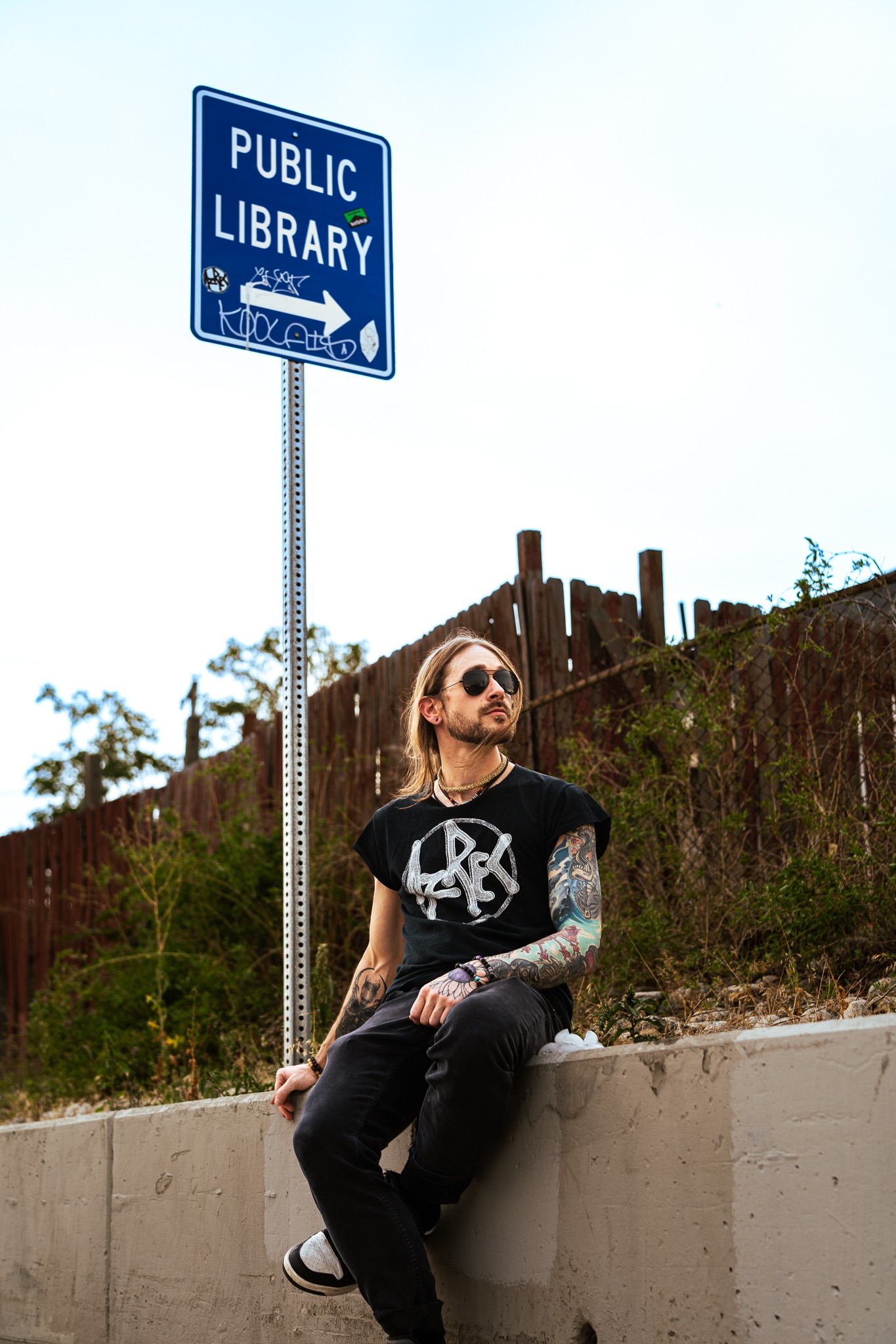Person with sunglasses and tattoos sitting on a concrete ledge near a public library sign, outdoors, with a fence and trees in background.