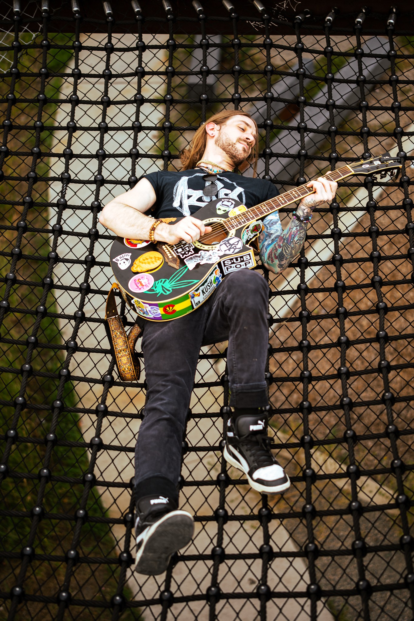 Person climbing a fence with a colorful guitar, head tilted back, outdoors, brick wall background.