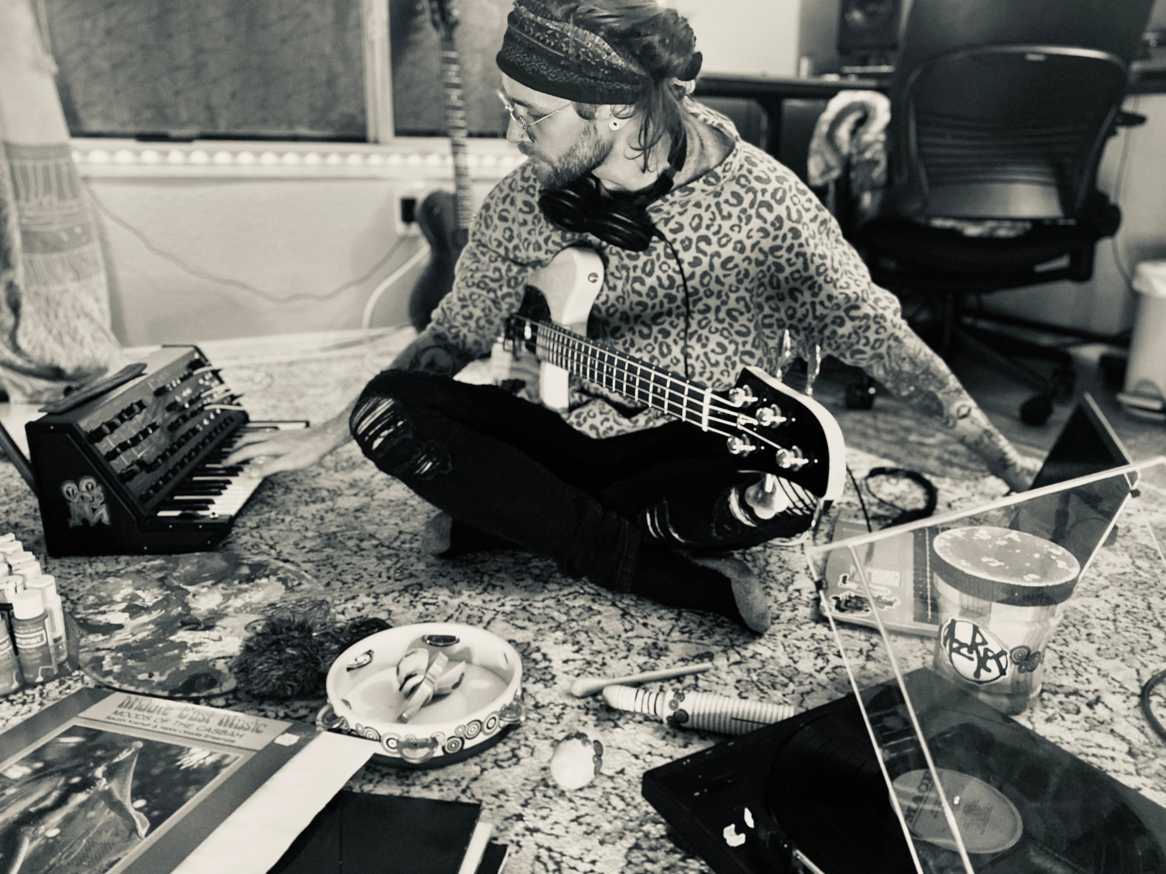 Person with long hair and glasses playing guitar at a cluttered table in a room, with a chair and window in background.