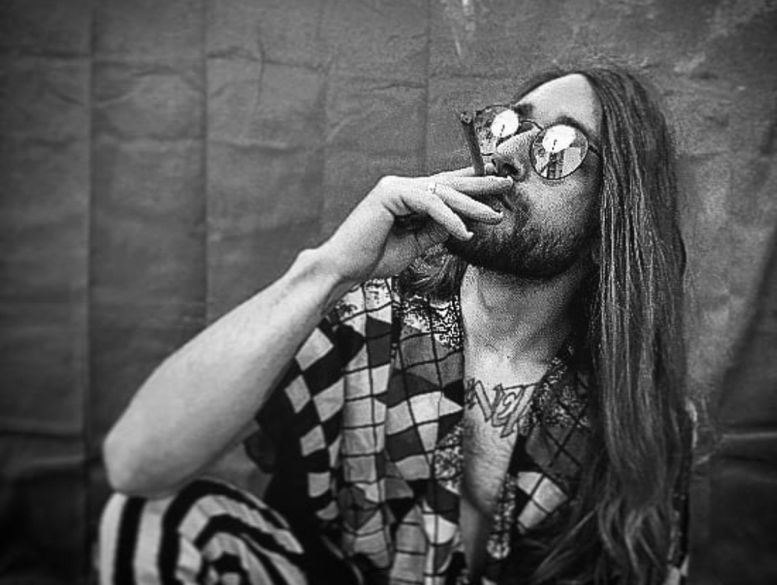 Person with long hair and sunglasses smokes a cigarette, sitting against a textured wall.