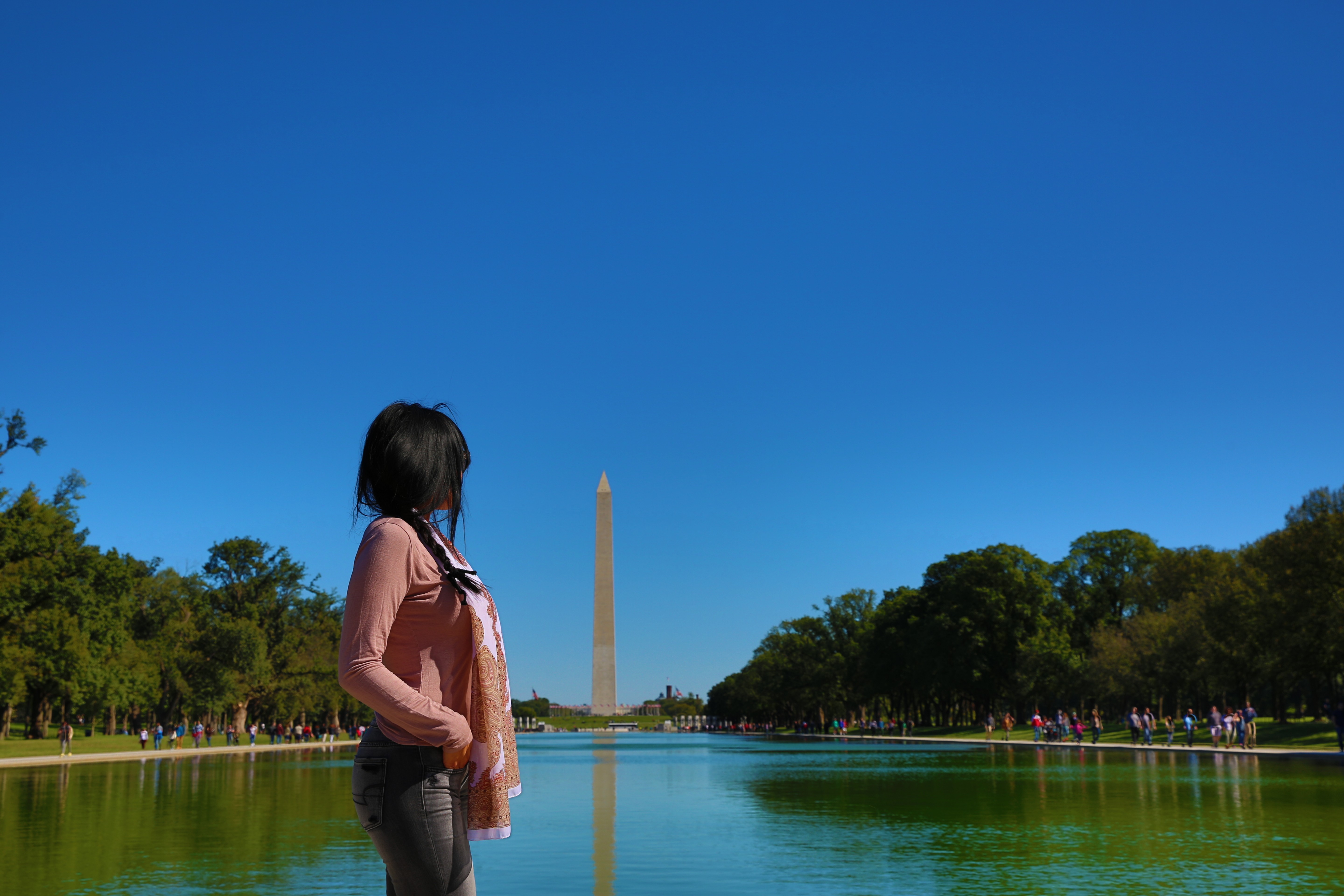 A person with long dark hair stands near a body of water with trees on both sides and a tall monument in the background under a clear blue sky.