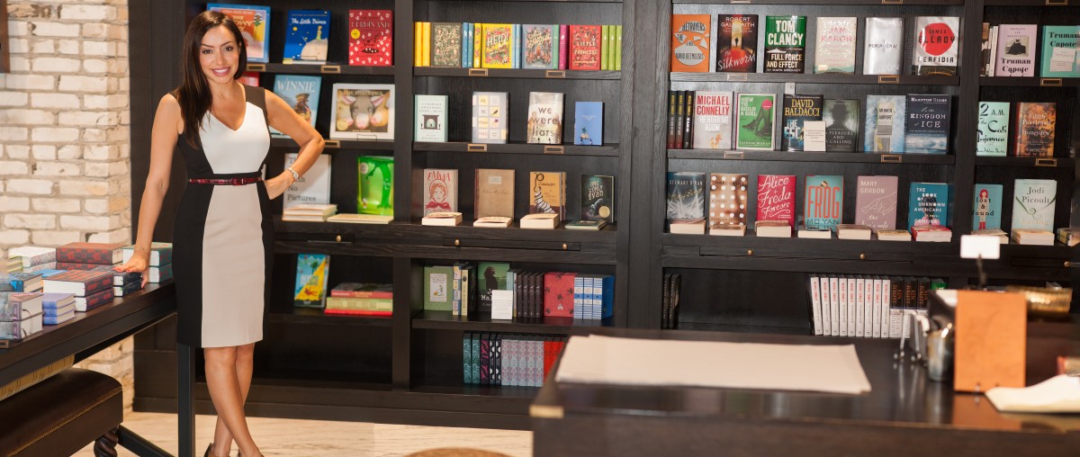 Woman standing in bookstore with shelves of books behind her, smiling, wearing a black and white dress.
