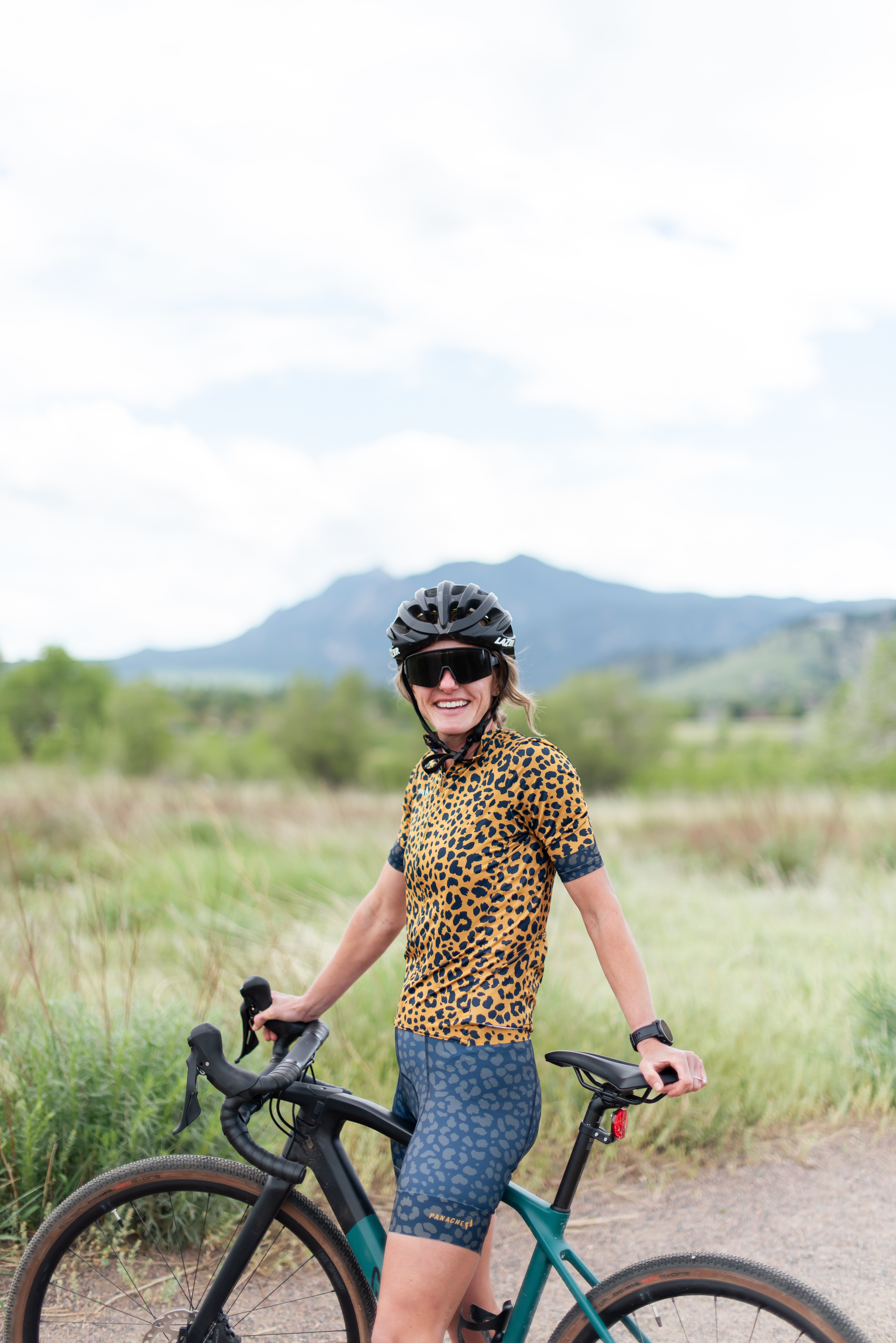 Child wearing a helmet and sunglasses, standing with a bicycle in a grassy field with mountains in the background.