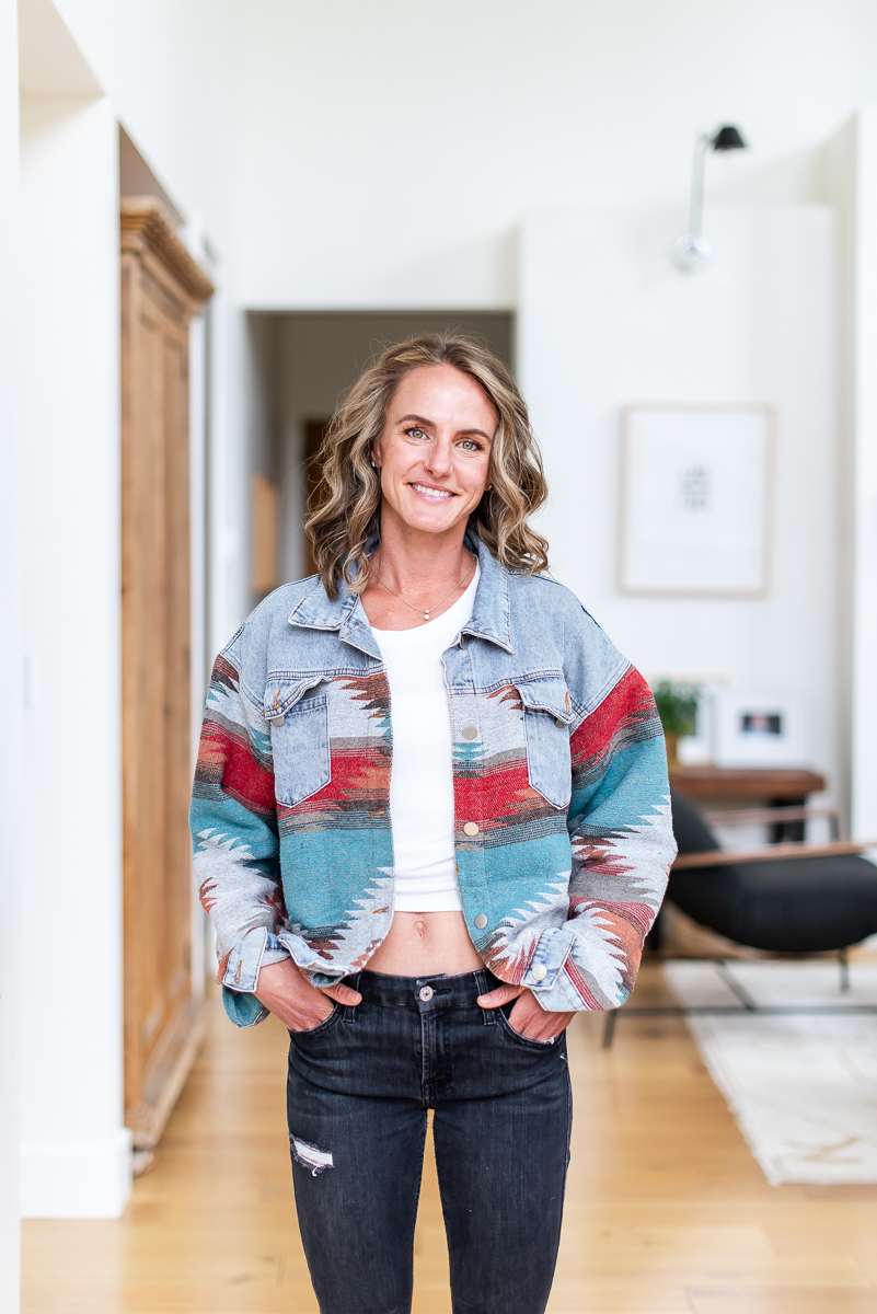 Woman with wavy hair smiling, wearing a colorful jacket and jeans, standing in a bright, modern room with wooden furniture.