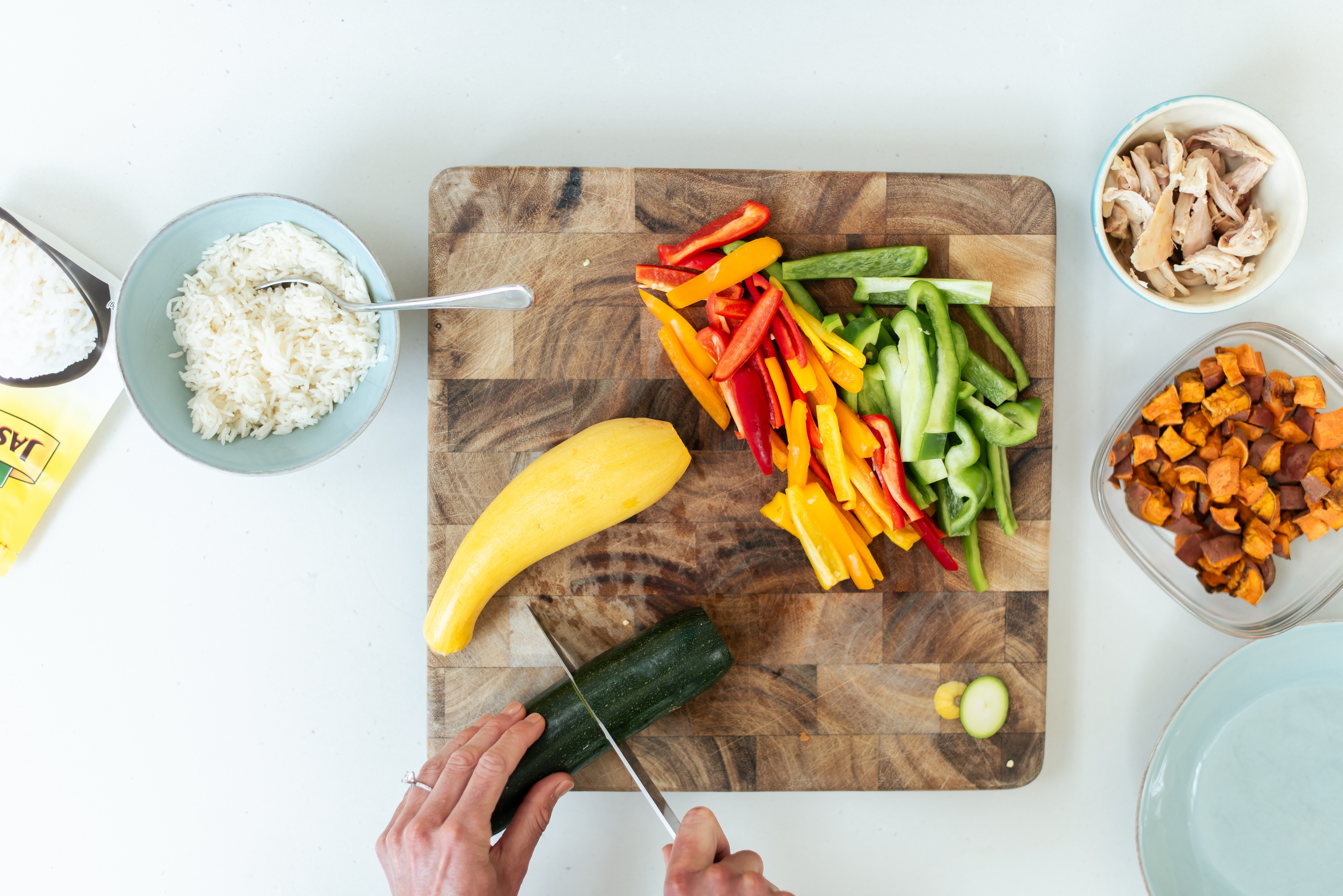 Person slicing cucumber on a wooden cutting board with sliced bell peppers, banana, and bowls of shredded chicken and diced vegetables nearby.