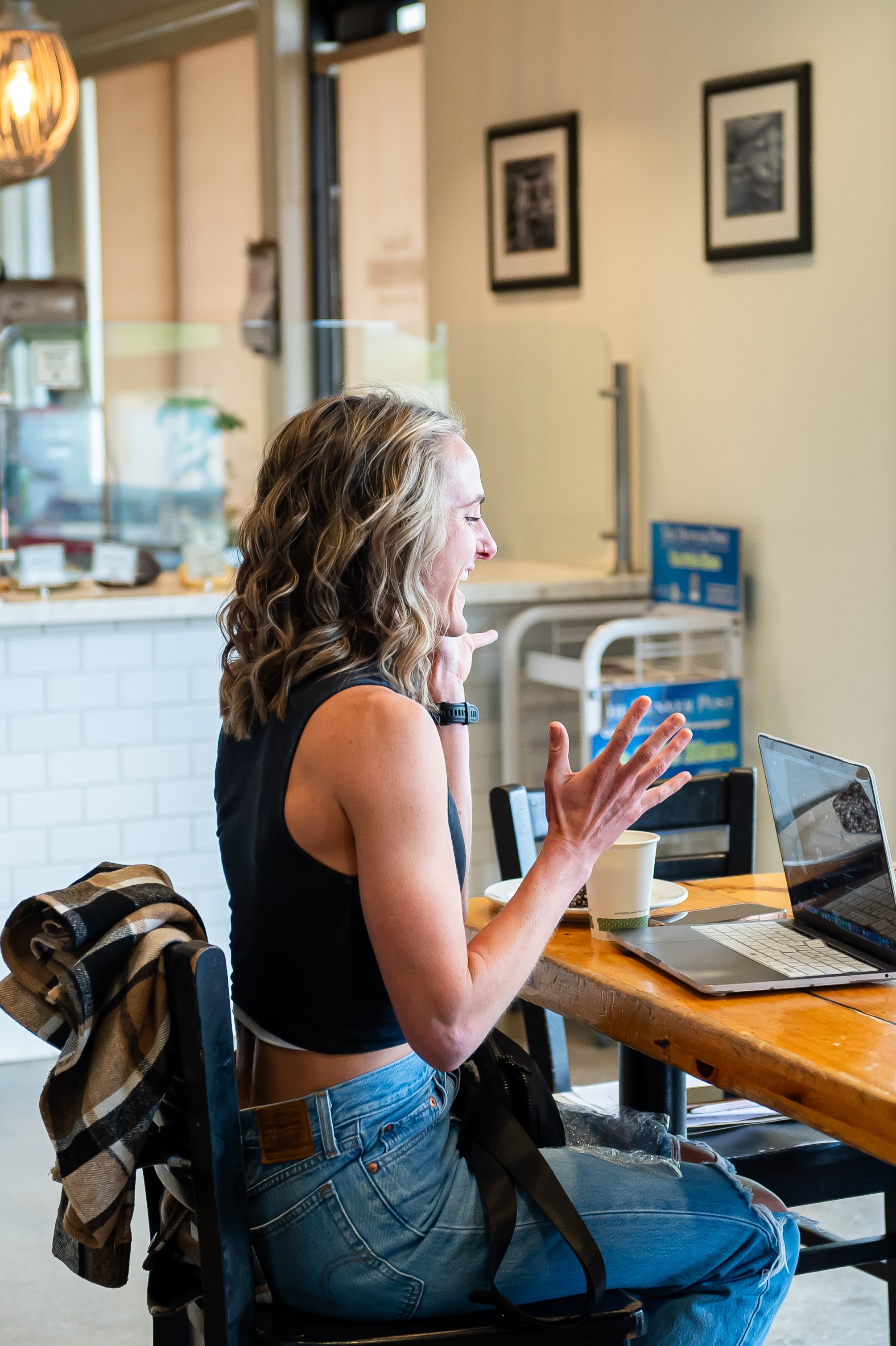 Woman with curly hair sitting at a table, gesturing with her hands, in a cafe setting, with laptops and a backpack nearby.