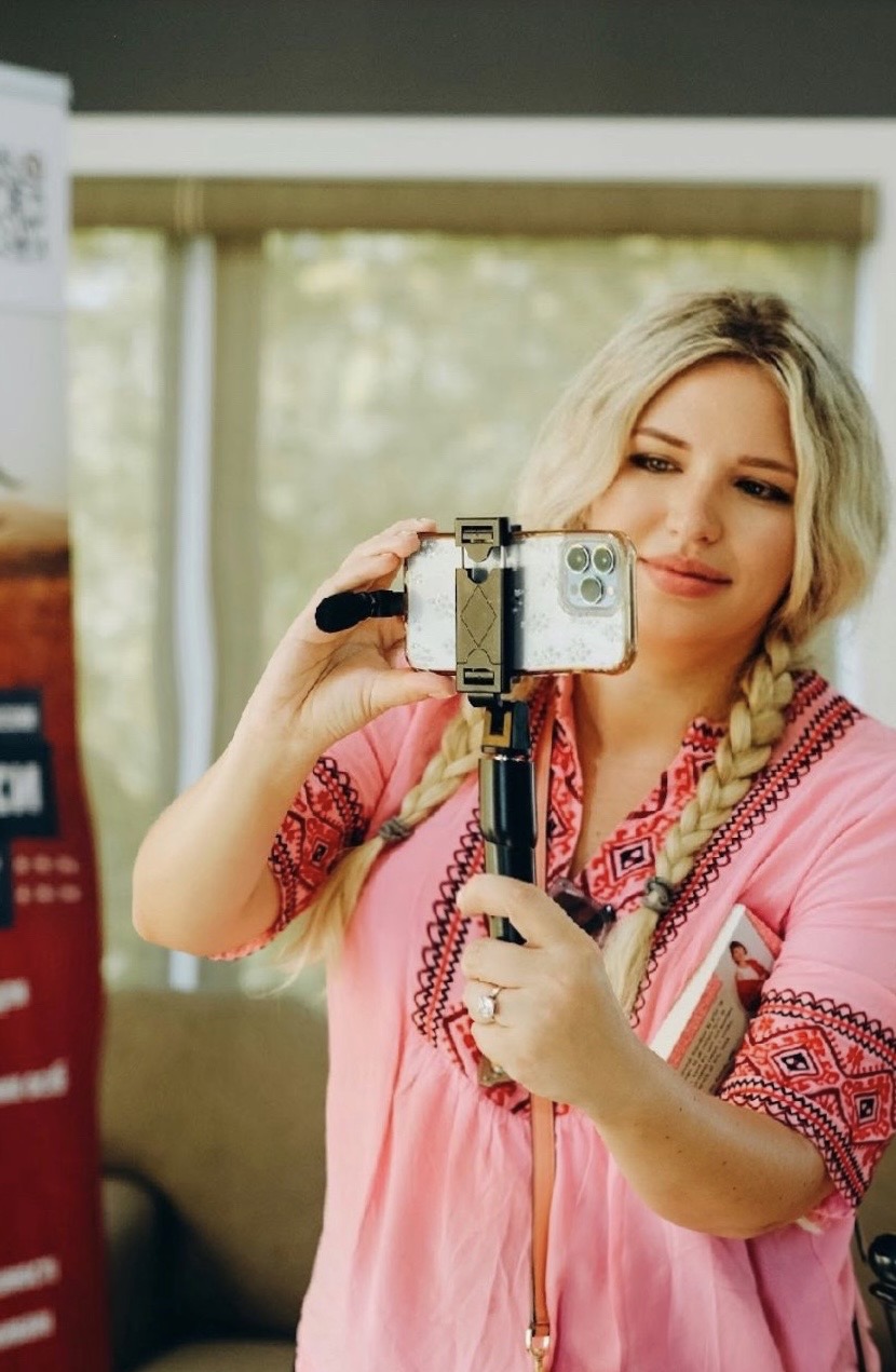 Woman with blonde hair in a braid taking a selfie with a camera, wearing a pink top with embroidery, indoors.