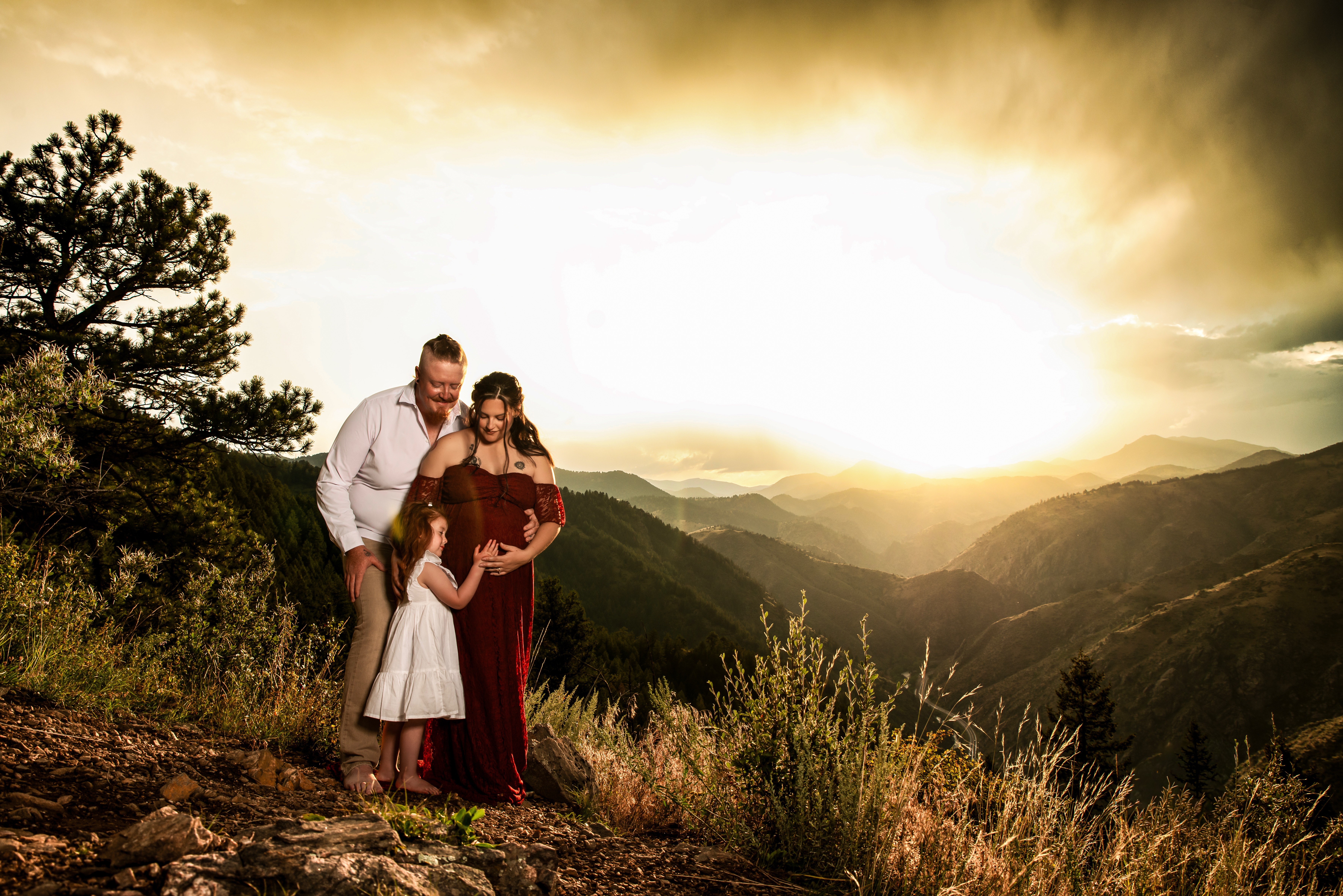 Family of three standing on a mountain trail during sunset with mountains and sky in background.