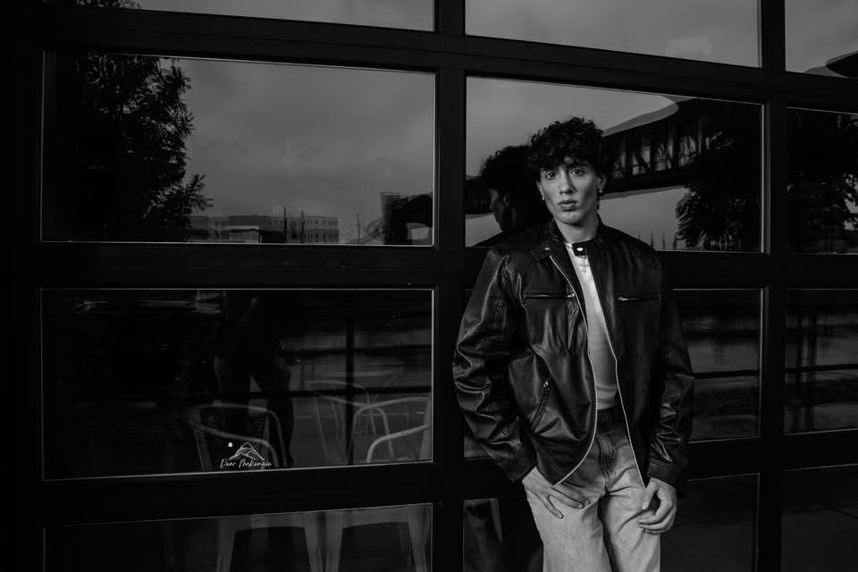 Young man with curly hair wearing a leather jacket standing in front of a large window with reflections, black and white photo.