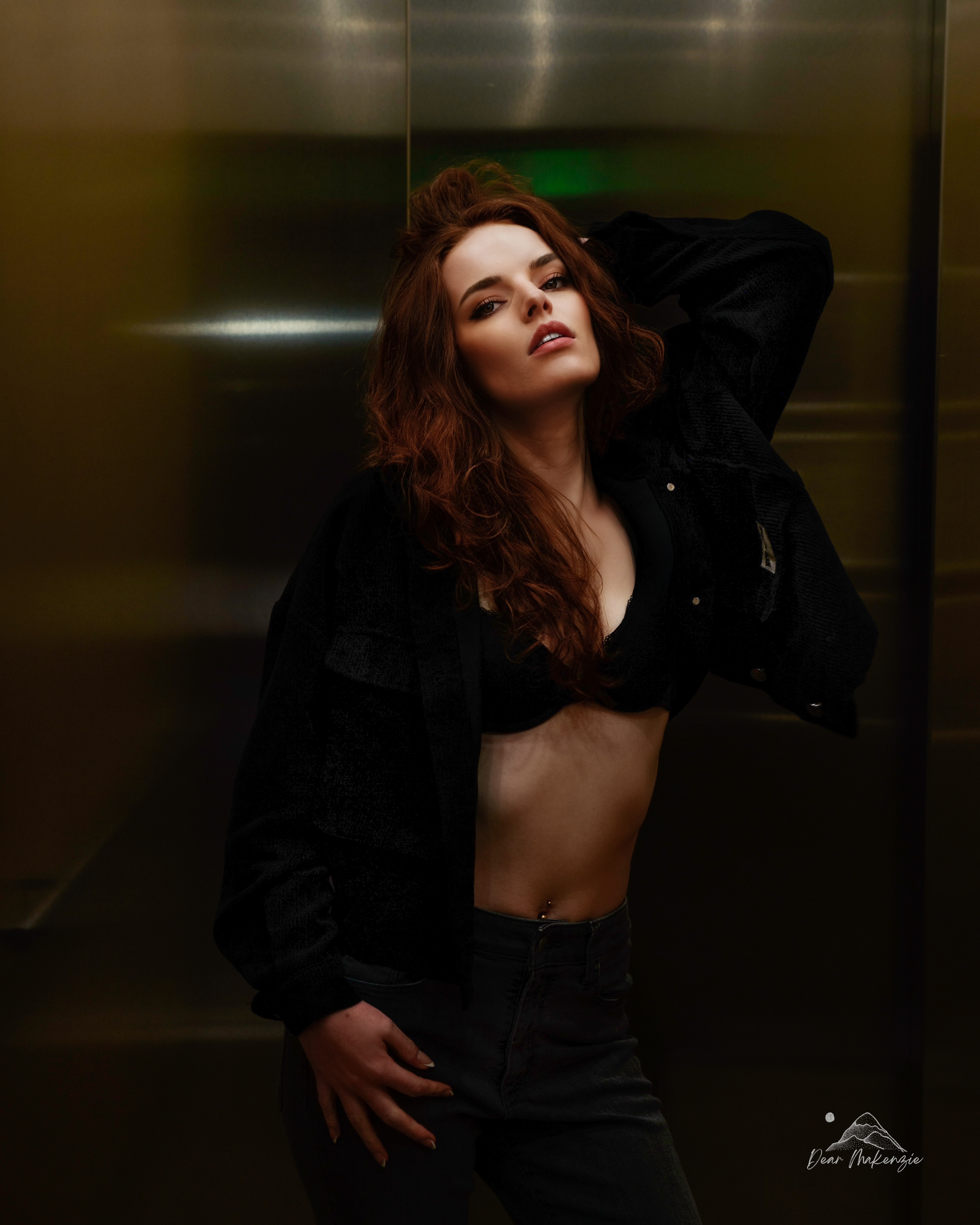 Young woman with long, wavy hair poses with one hand behind her head, wearing a black jacket and crop top, in an elevator.